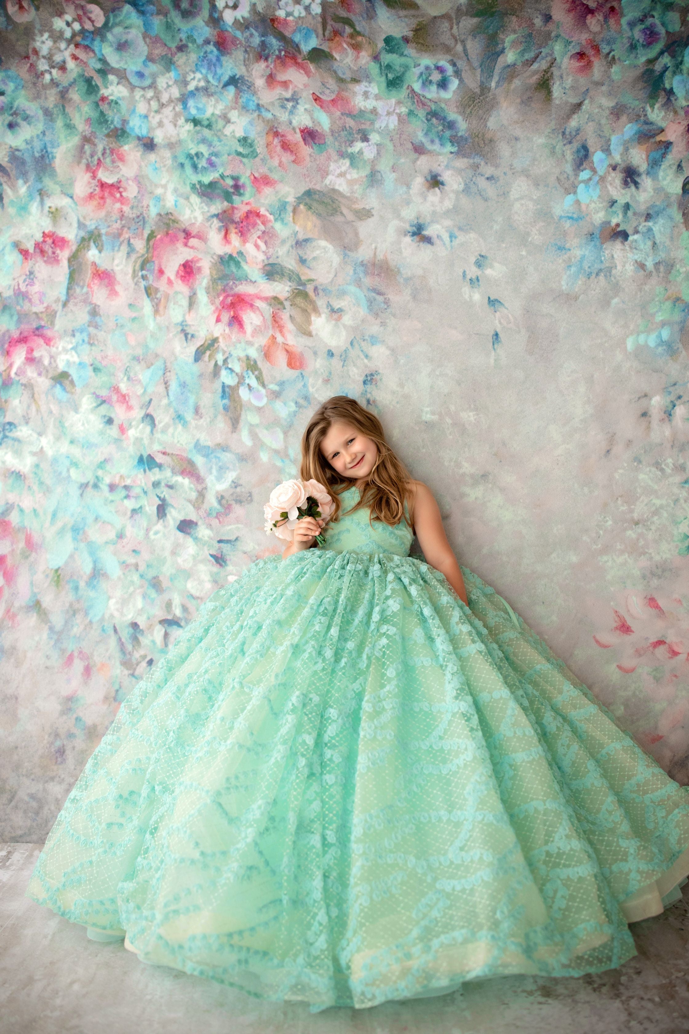 A young girl wearing a floor-length mint green gown with a floral pattern, standing in front of a colorful floral backdrop.