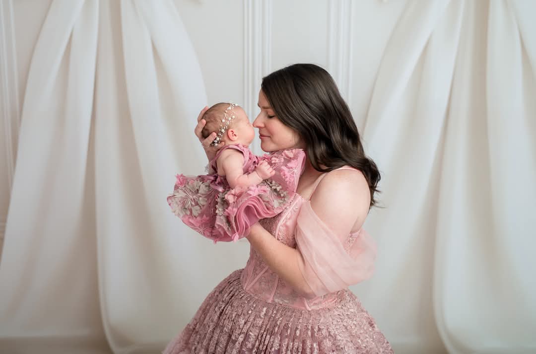 Woman holding a baby dressed in pink against a white curtain background