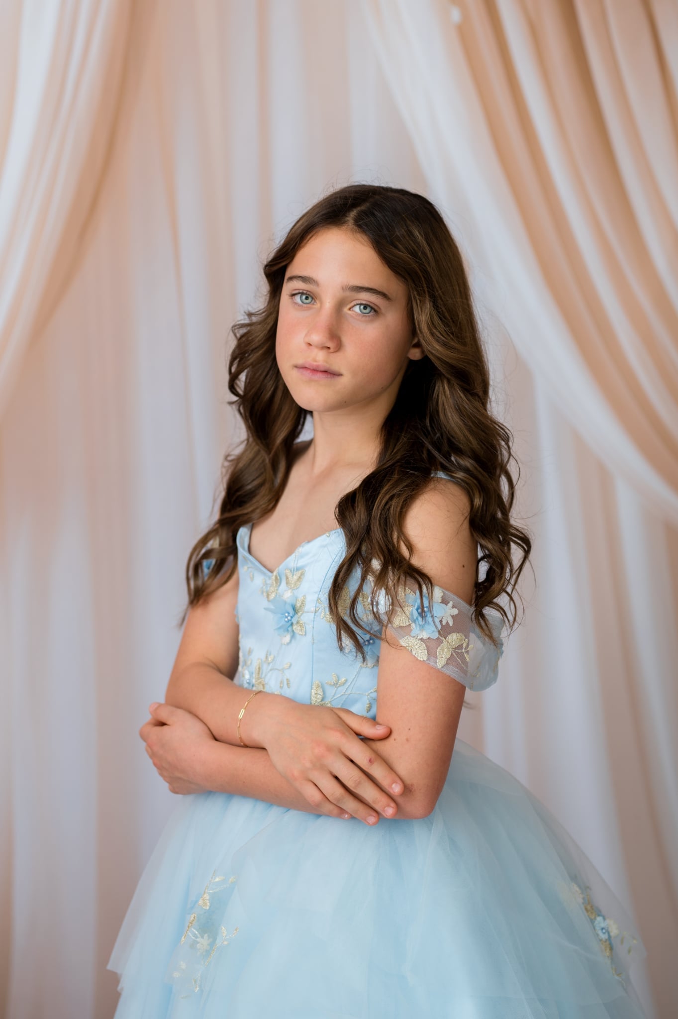 Young girl in a light blue dress with floral details against a soft pink curtain background
