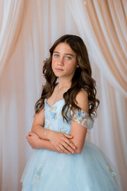 Young girl in a light blue dress with floral details against a soft pink curtain background
