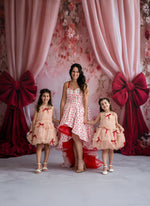 Woman with two young girls in matching dresses standing in front of pink floral curtains with red bows.