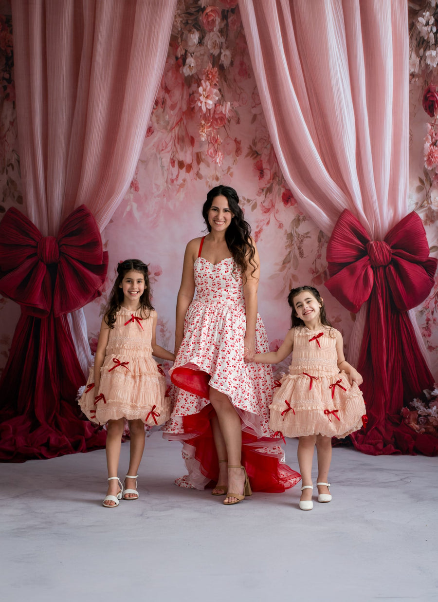 Woman with two young girls in matching dresses standing in front of pink floral curtains with red bows.