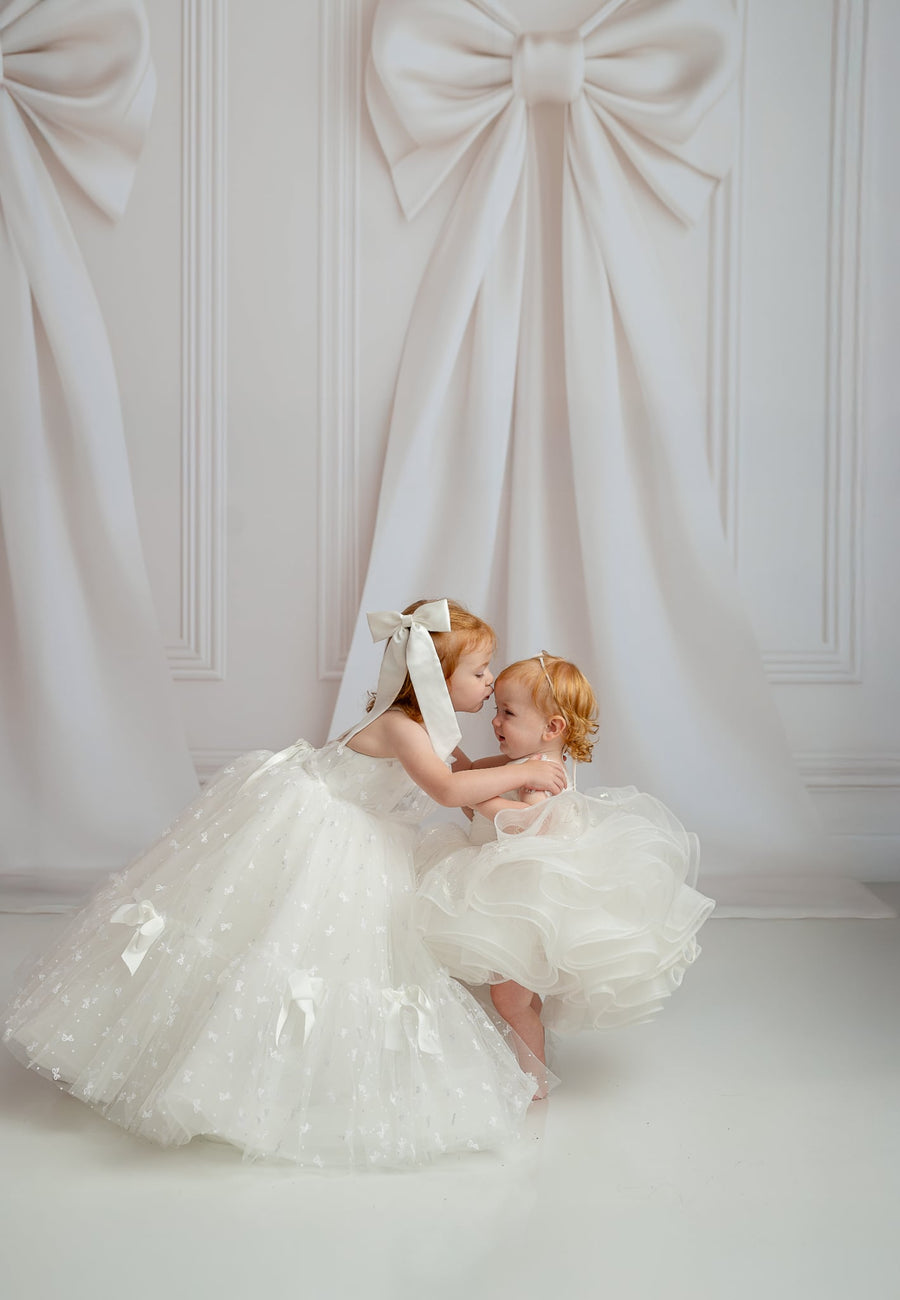 Two young girls in white dresses with large bows against a white wall.