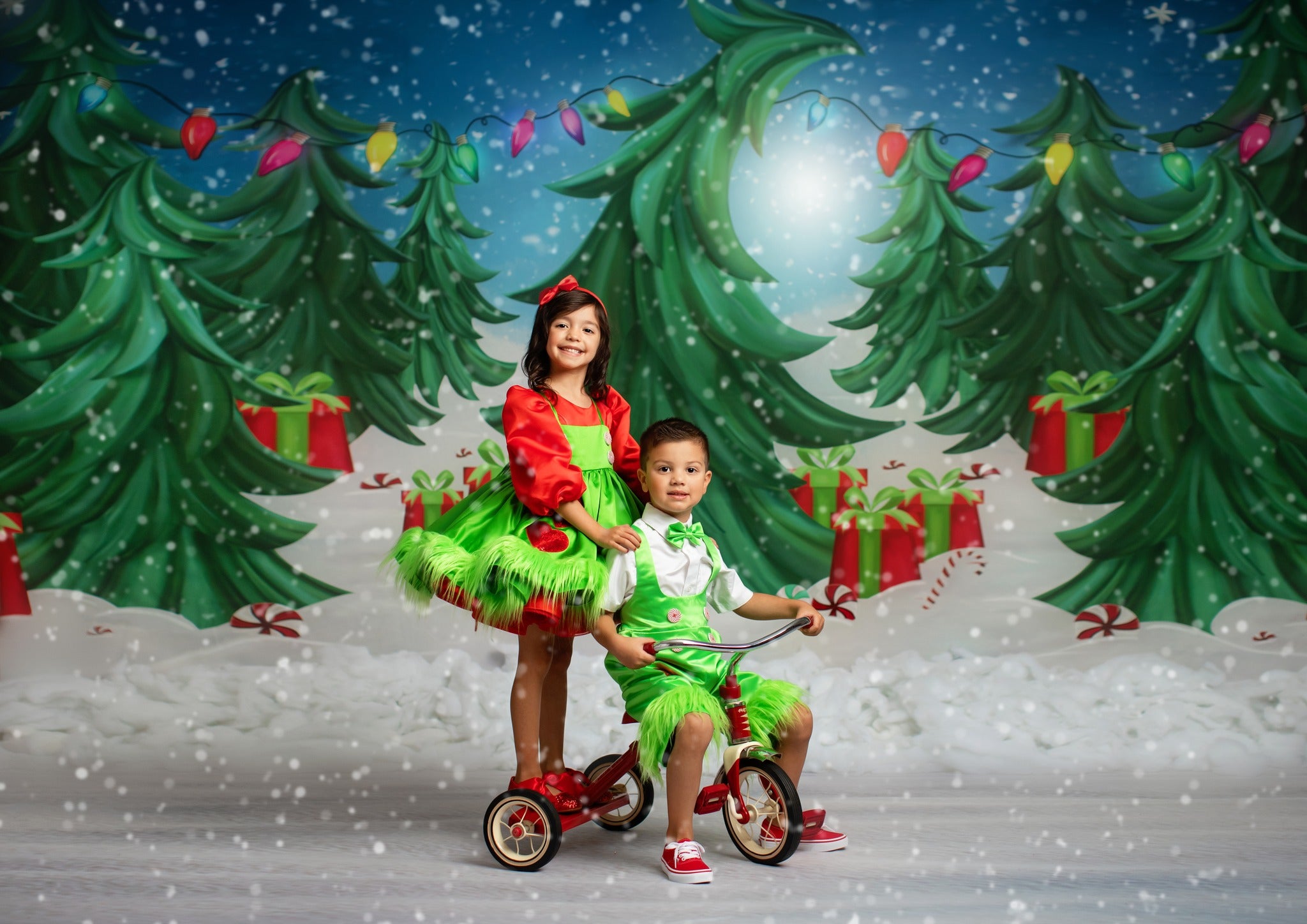 Two children dressed in vibrant green and red Christmas-themed outfits, smiling in front of a festive holiday backdrop with decorated trees and gifts.
