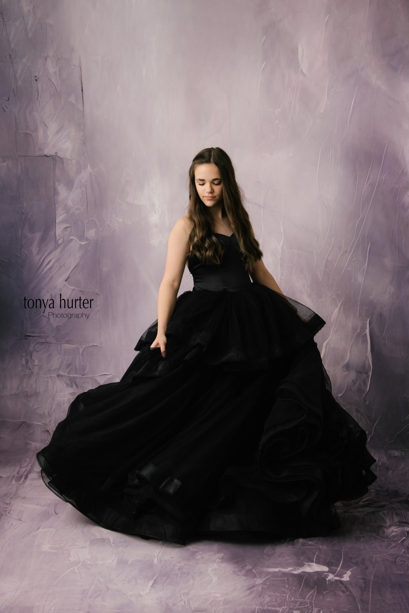 "A young girl posing in an elegant black gown with a sweetheart neckline and shimmering fabric that catches the light."
