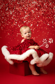 Child in a red dress with white fur trim sitting on a red box against a red background with candy cane decorations.