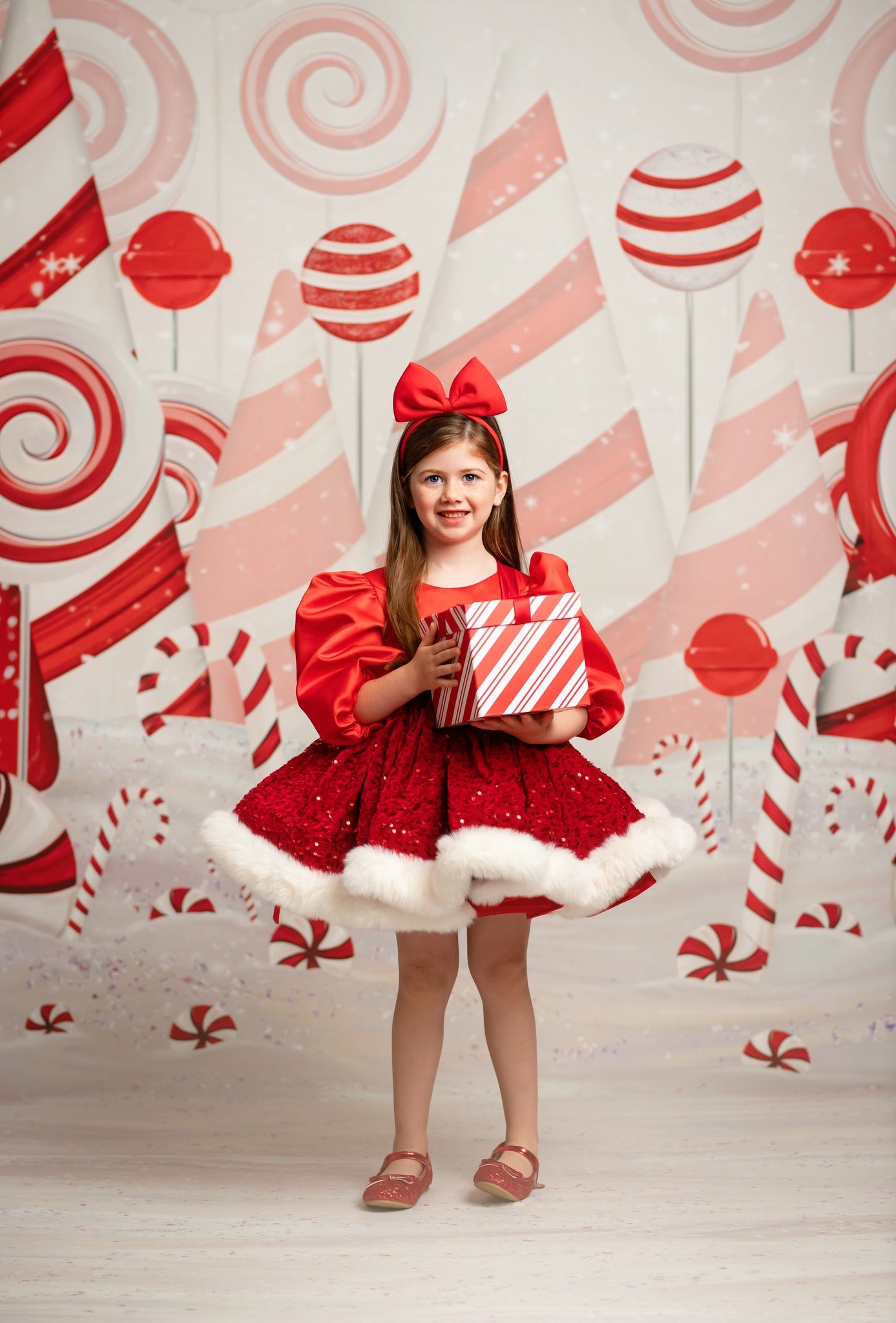 Young girl in a red dress with white fur trim holding a gift box against a candy-themed backdrop.