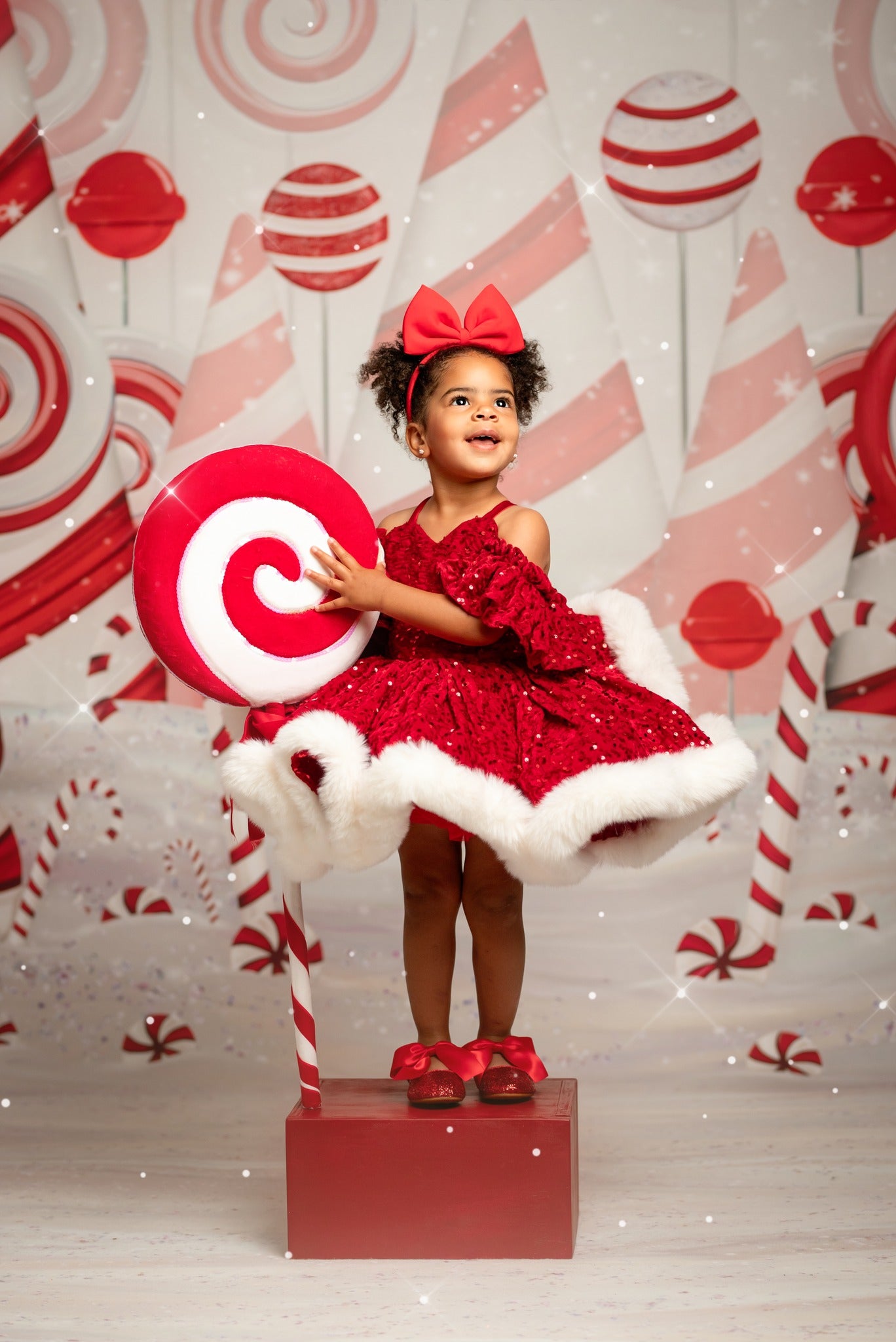 Young girl in a red dress with white fur trim and a red bow, holding a candy cane, against a festive background with candy-themed decorations.