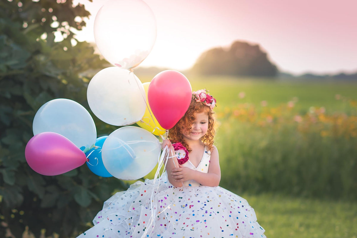 Child dressed in a whimsical white gown with colorful dots, posing with balloons on a grassy field.
