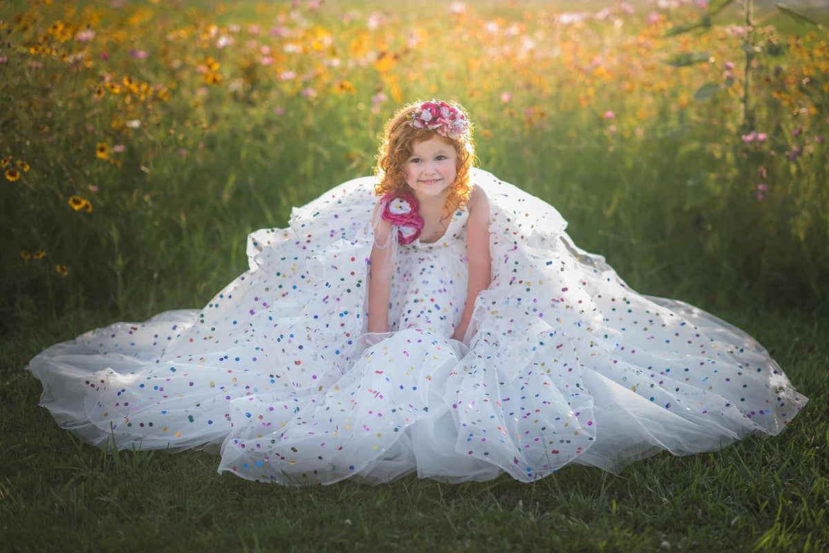 Little girl wearing a confetti-covered gown with floral shoulder accents, smiling in a field at sunset.
