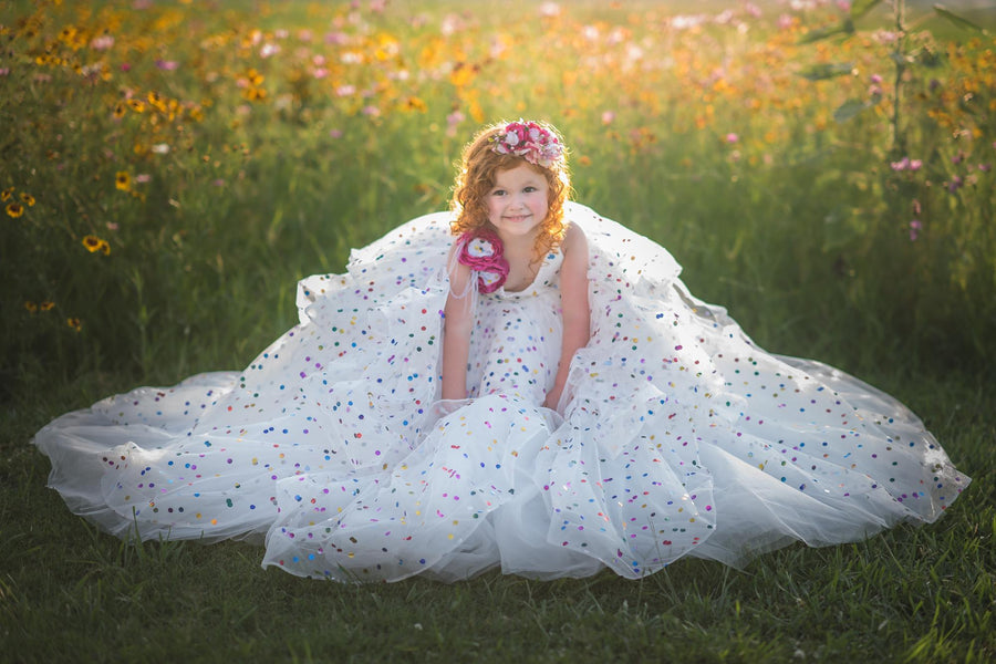 Little girl wearing a confetti-covered gown with floral shoulder accents, smiling in a field at sunset.
