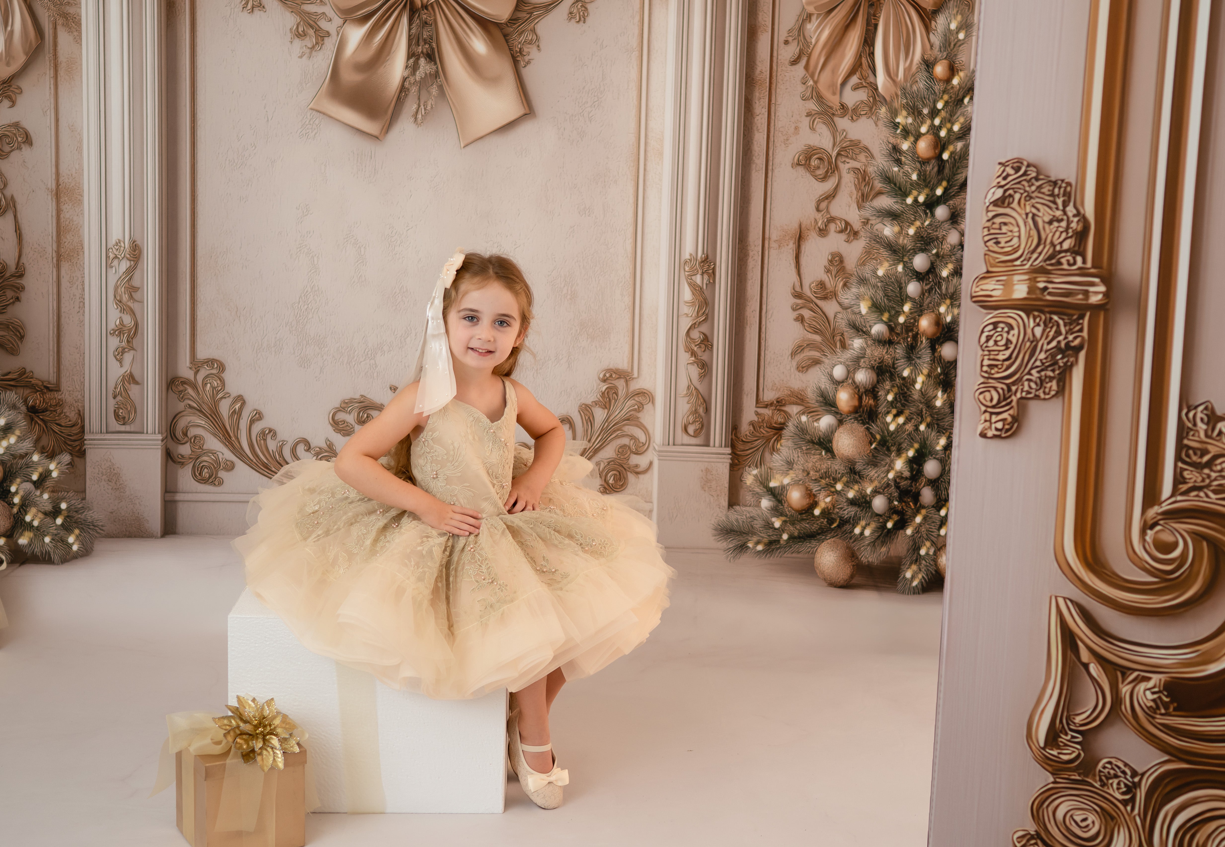 Young girl in a beige dress sitting on a white box in a decorated room with Christmas trees.