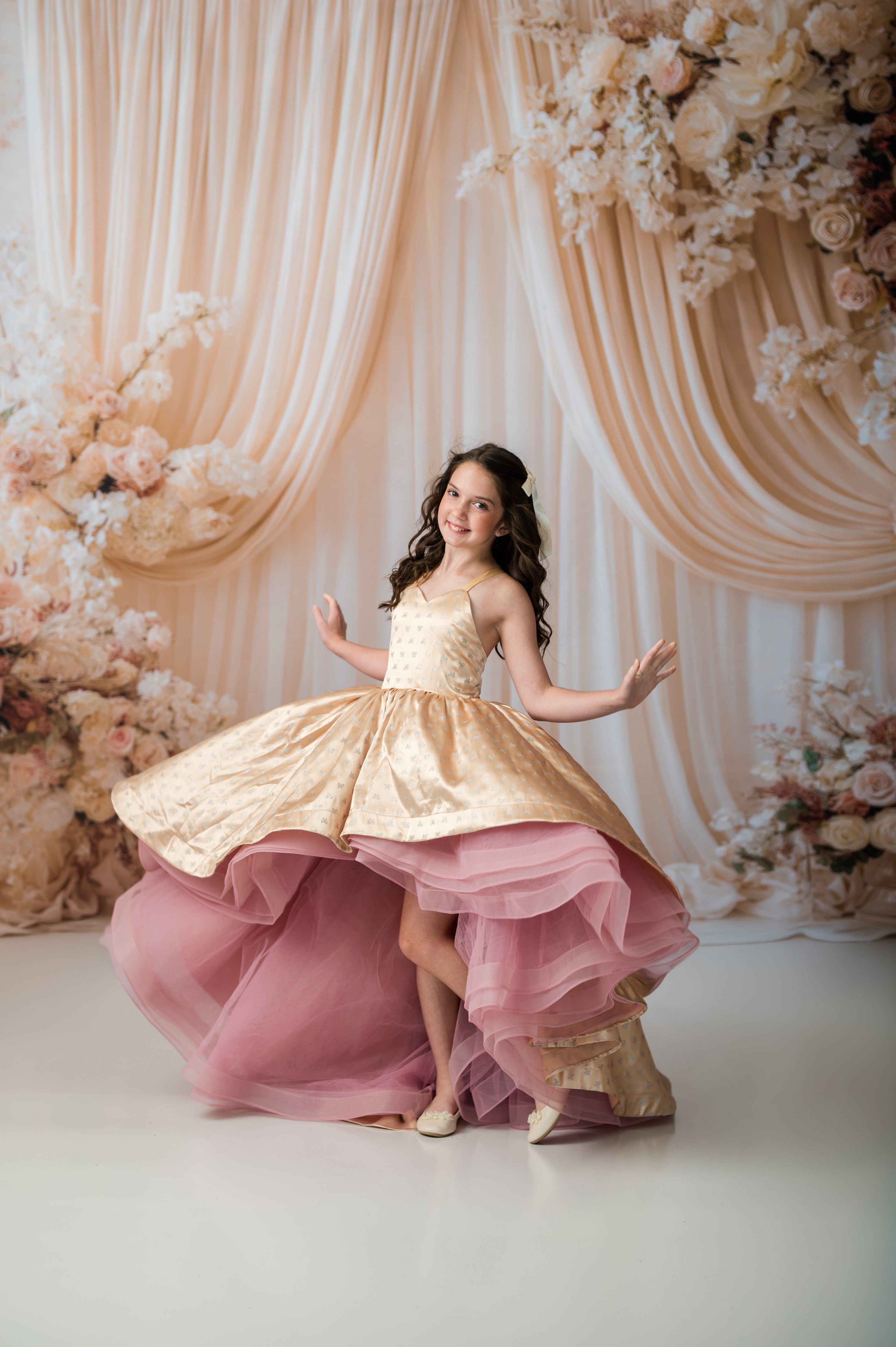 A young girl with long dark hair poses in a formal ballgown with a gold satin bodice and a layered pink tulle skirt, set against a backdrop of draped cream fabric and floral arrangements.