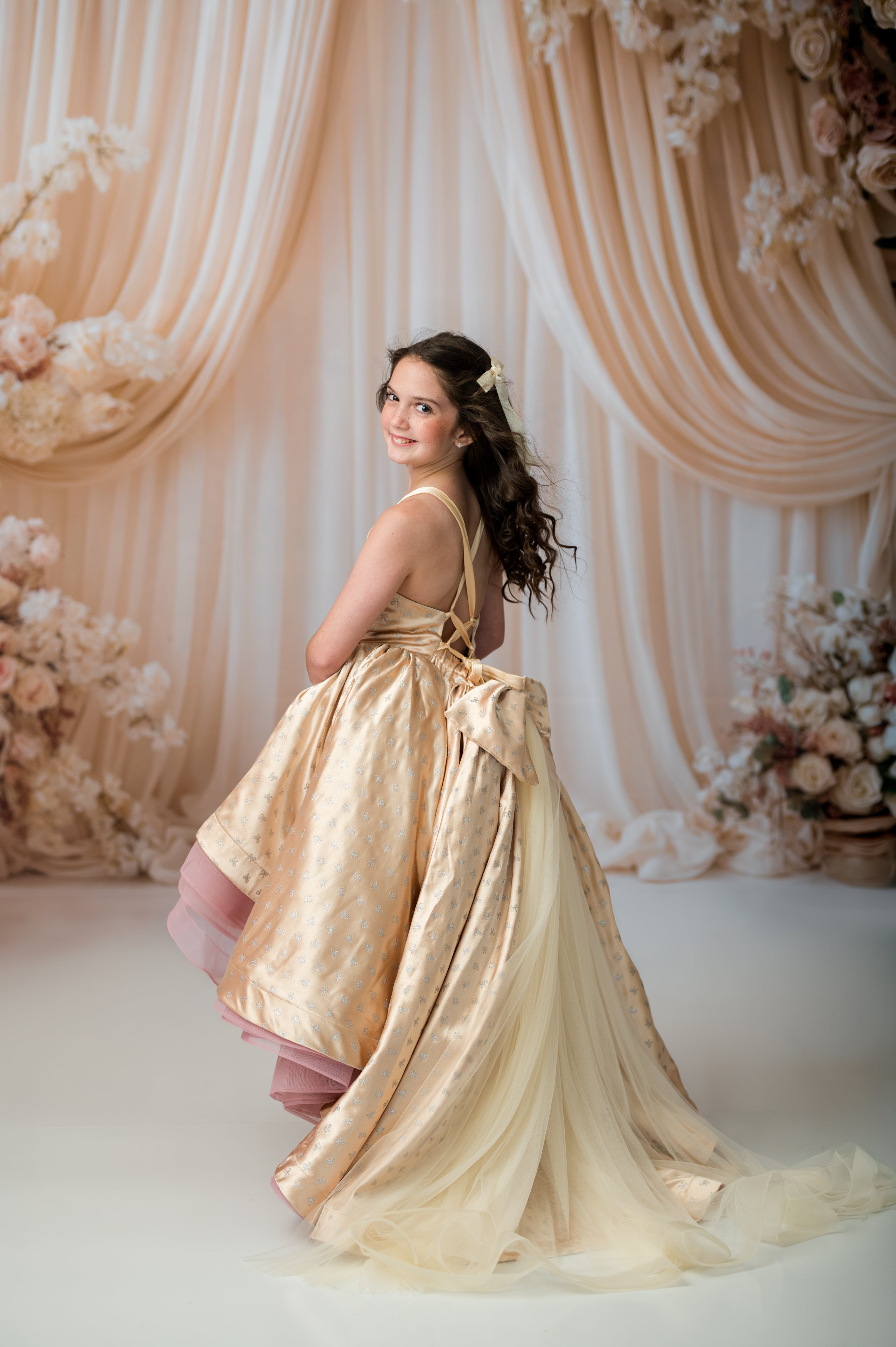 A young woman wearing a formal ball gown with a large bow at the back, posed in front of a draped beige fabric backdrop with floral arrangements.