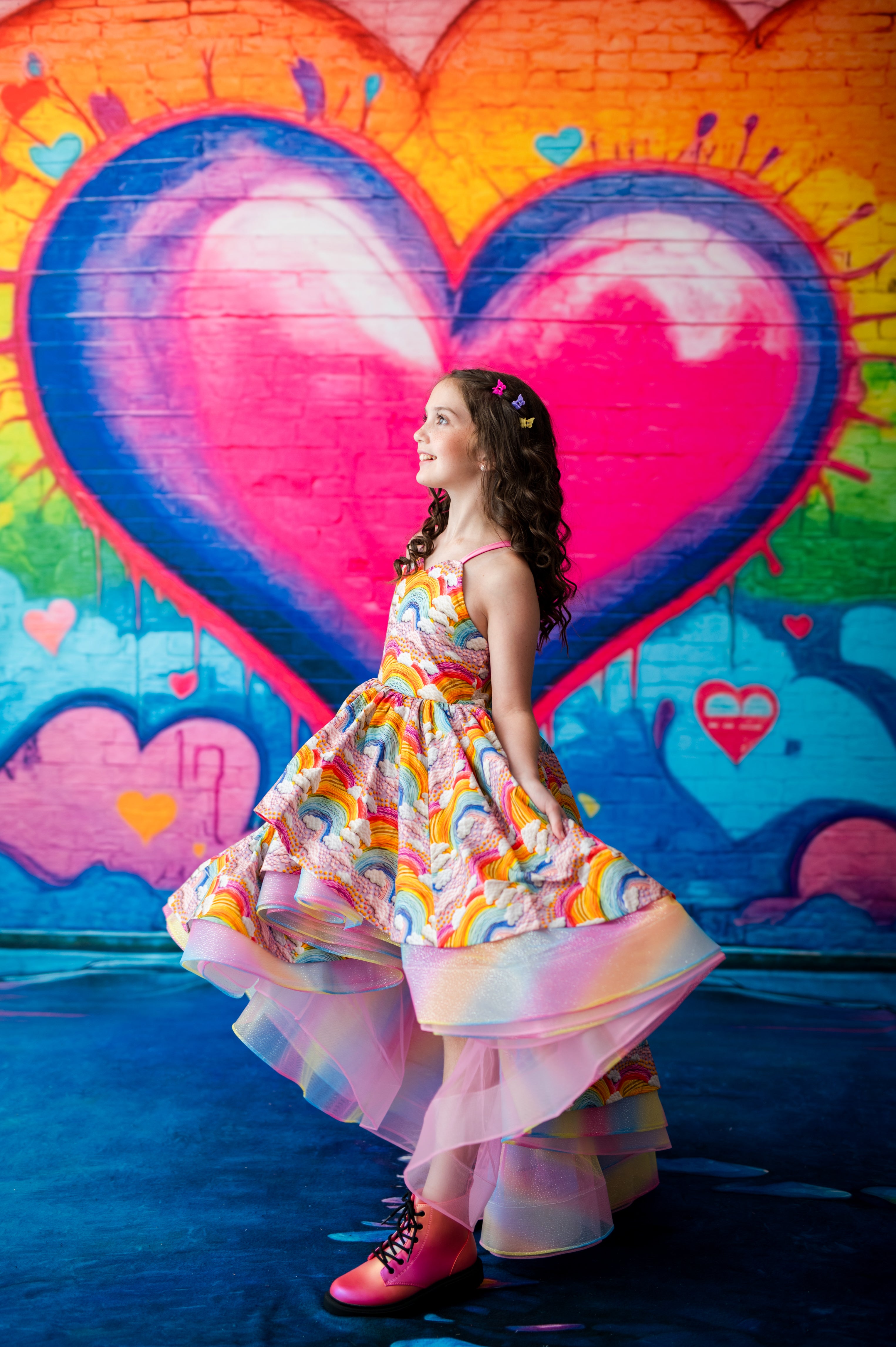 A young girl with curly brown hair is wearing a colorful, rainbow-patterned tiered dress and pink lace-up boots, standing in front of a large, vibrant graffiti-style heart mural.