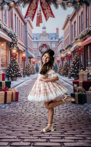 A young girl posing in a festive holiday street scene, wearing a white tulle dress with a peach floral pattern and white block-heel sandals. The background is a decorated Christmas street with trees, gifts, and garlands.
