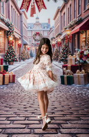 A young girl with long dark hair stands on a cobblestone street wearing a white tulle dress with a beige floral pattern and white heels. The background depicts a festive Christmas town scene with decorated trees, gift boxes, and red awnings.