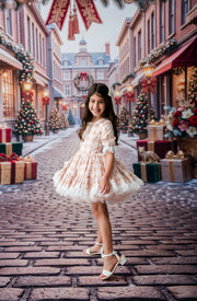 A young girl stands on a cobblestone street wearing a short, ruffled cream dress with a delicate pink floral pattern and white tulle trim. The background is a festive Christmas town scene with decorated trees, gift boxes, and red banners.