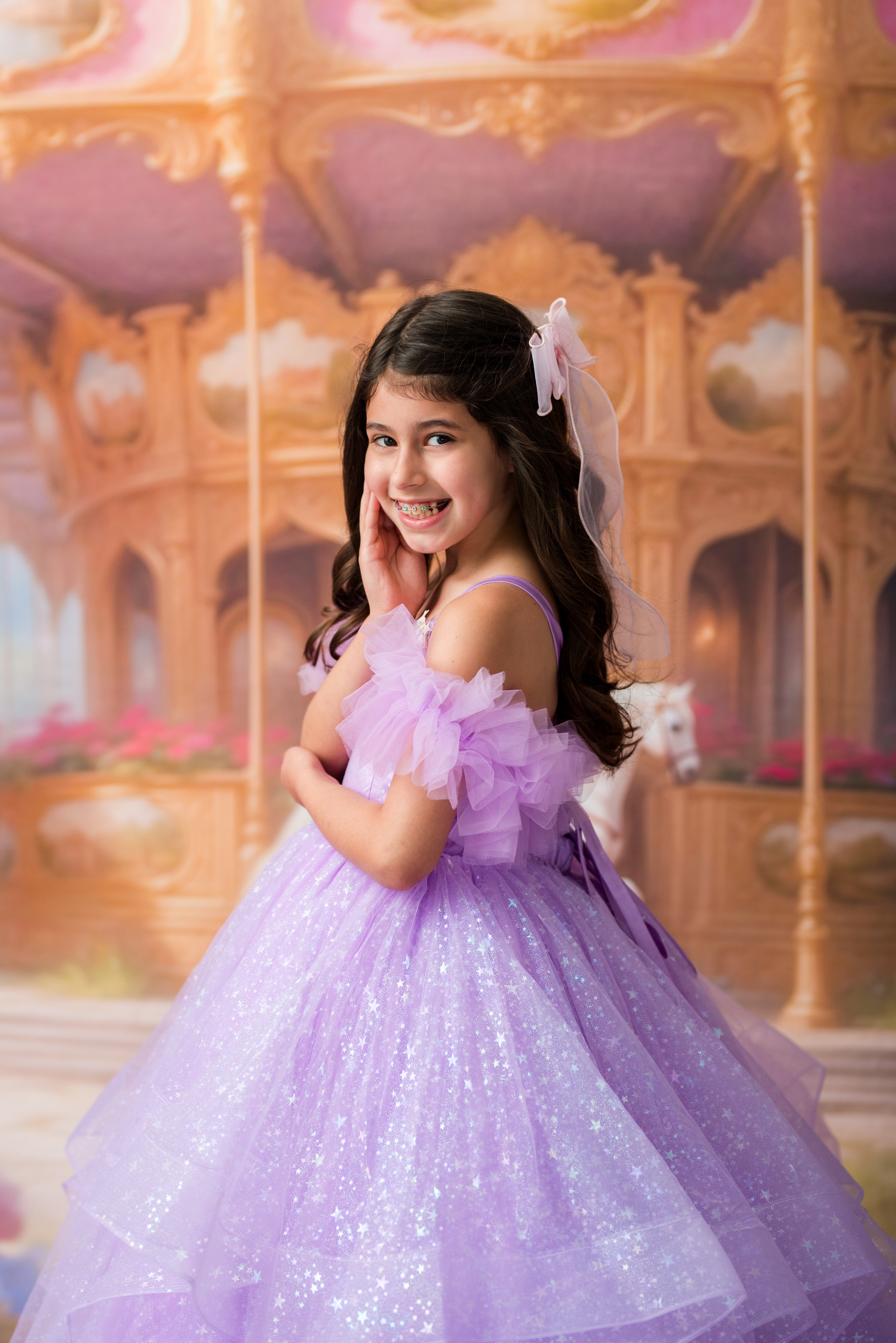 A young girl with long dark hair wearing a sparkling lavender ball gown with ruffled off-the-shoulder sleeves and a matching hair bow, posing in front of a golden carousel backdrop.