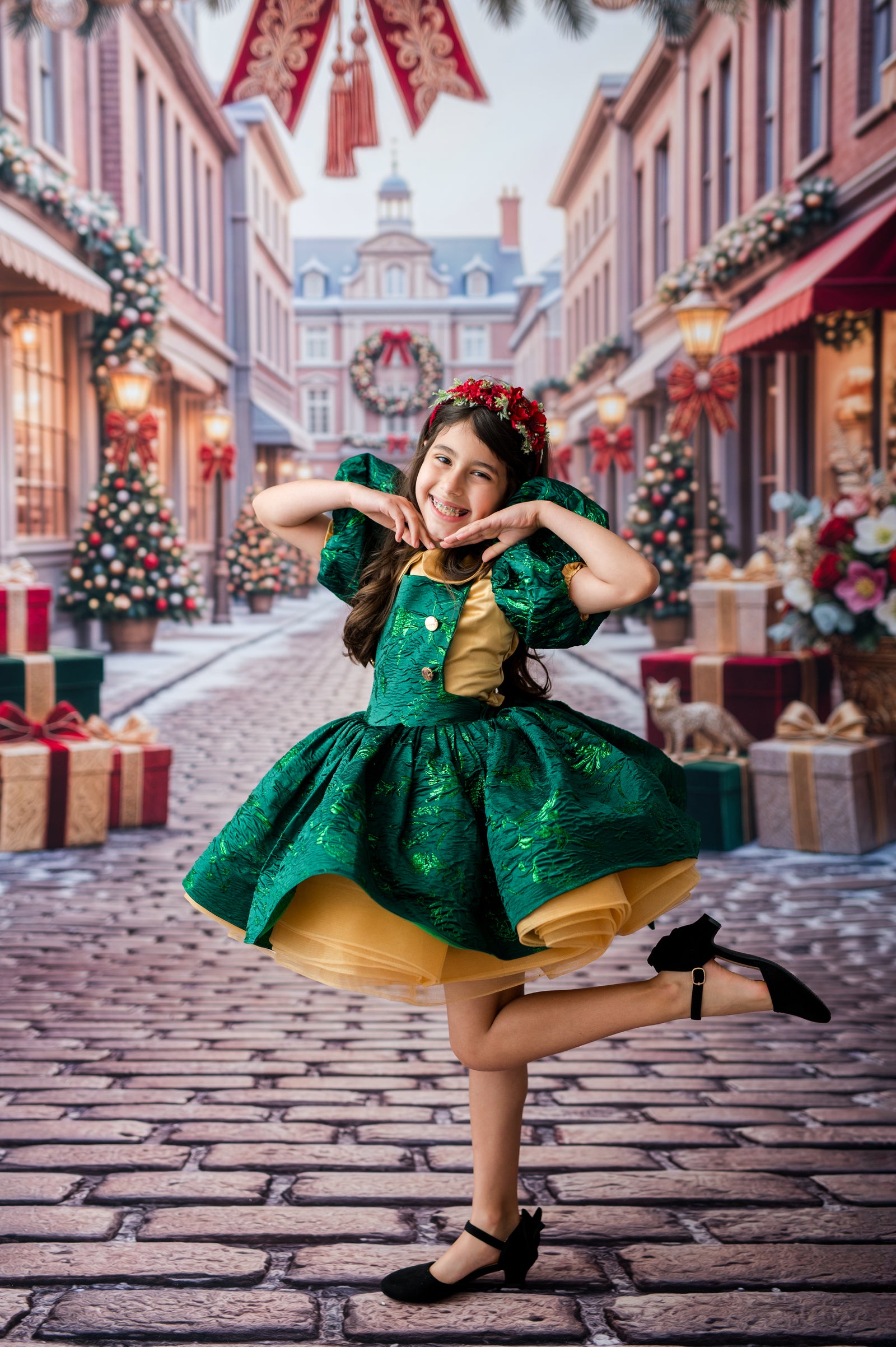 A young girl posing in a festive holiday street scene, wearing a green and gold dress with black heels. The background shows a beautifully decorated cobblestone street with Christmas trees, gift boxes, and warm lights.