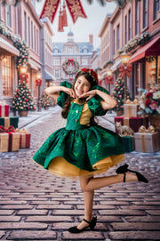 A young girl posing in a festive holiday street scene, wearing a green and gold dress with black heels. The background shows a beautifully decorated cobblestone street with Christmas trees, gift boxes, and warm lights.