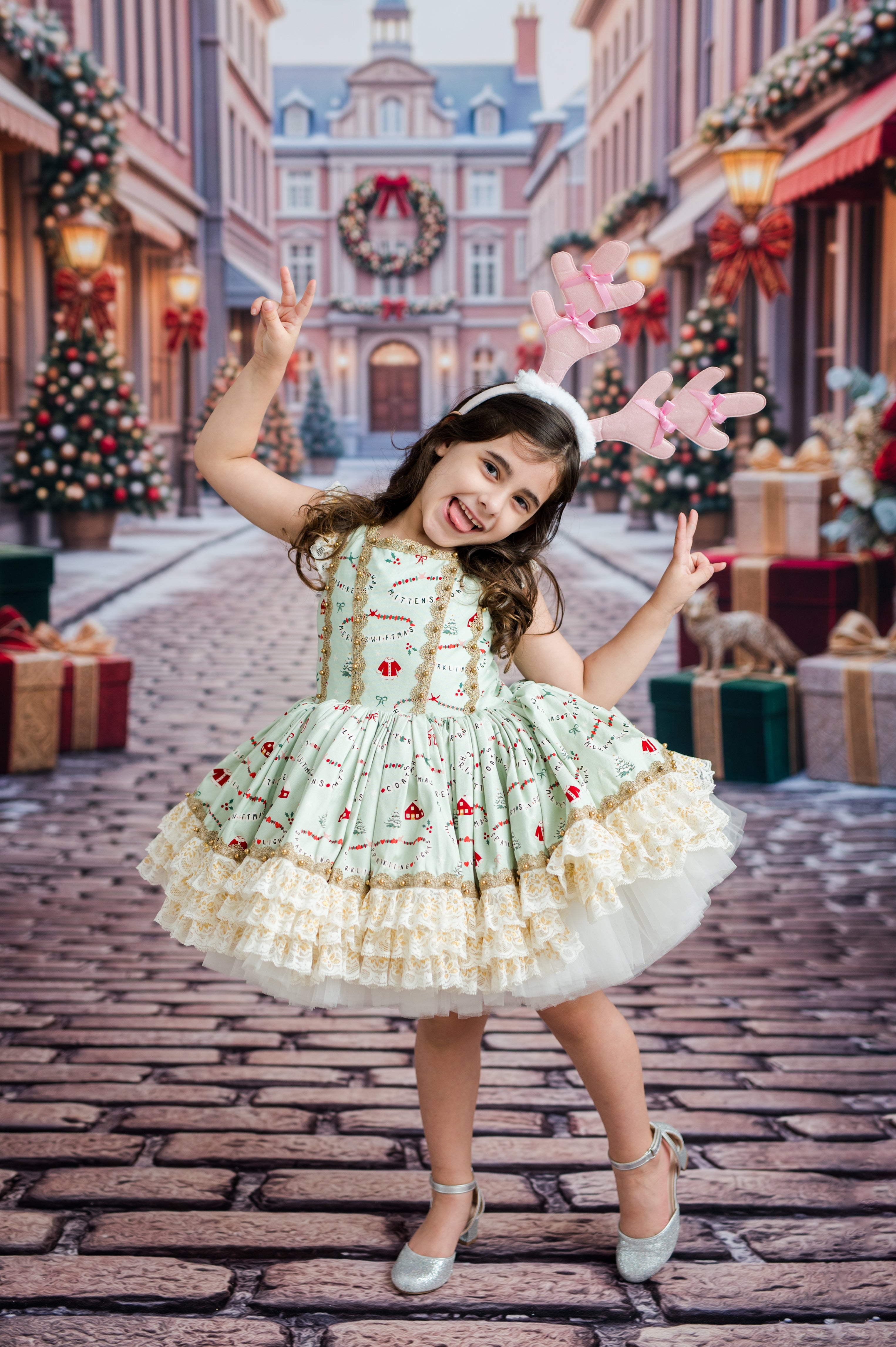 A young girl posing in a festive holiday street scene, wearing a light green and cream tiered dress with red berry prints, pink reindeer antler headband, and silver shoes.