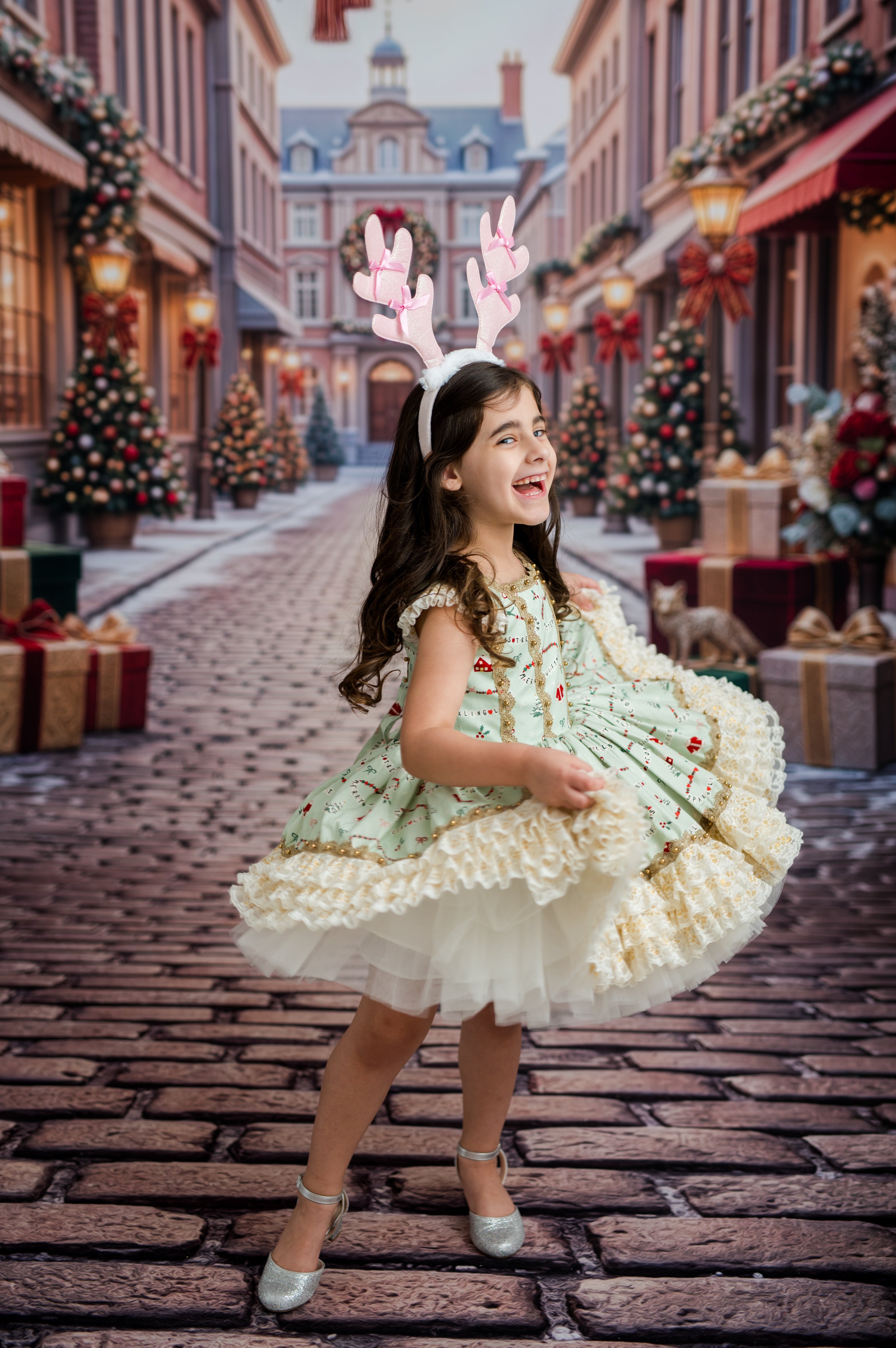 A young girl wearing a festive light green and cream tulle dress with red berry embroidery, pink bunny antlers, and silver shoes, posing on a cobblestone street decorated for Christmas.