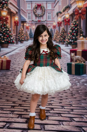 A young girl stands in a festive Christmas town street wearing a holiday dress with a green and red plaid bodice, white ruffled skirt, brown boots, and white socks. The background shows decorated Christmas trees, wreaths, and gift boxes.