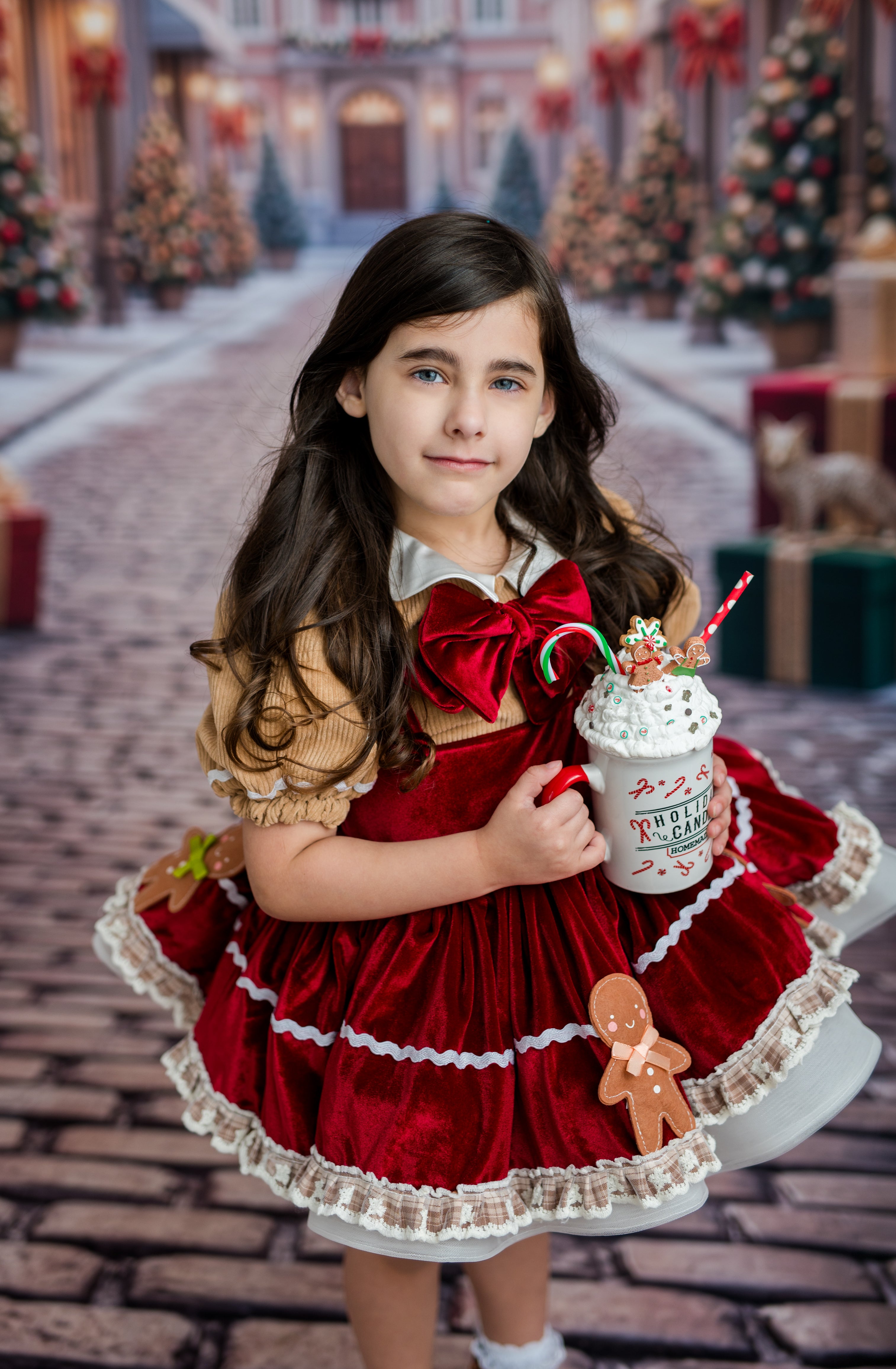 A young girl with long dark hair stands in a festive holiday village scene, wearing a red and beige dress with white lace trim and holding a decorated holiday drink cup.