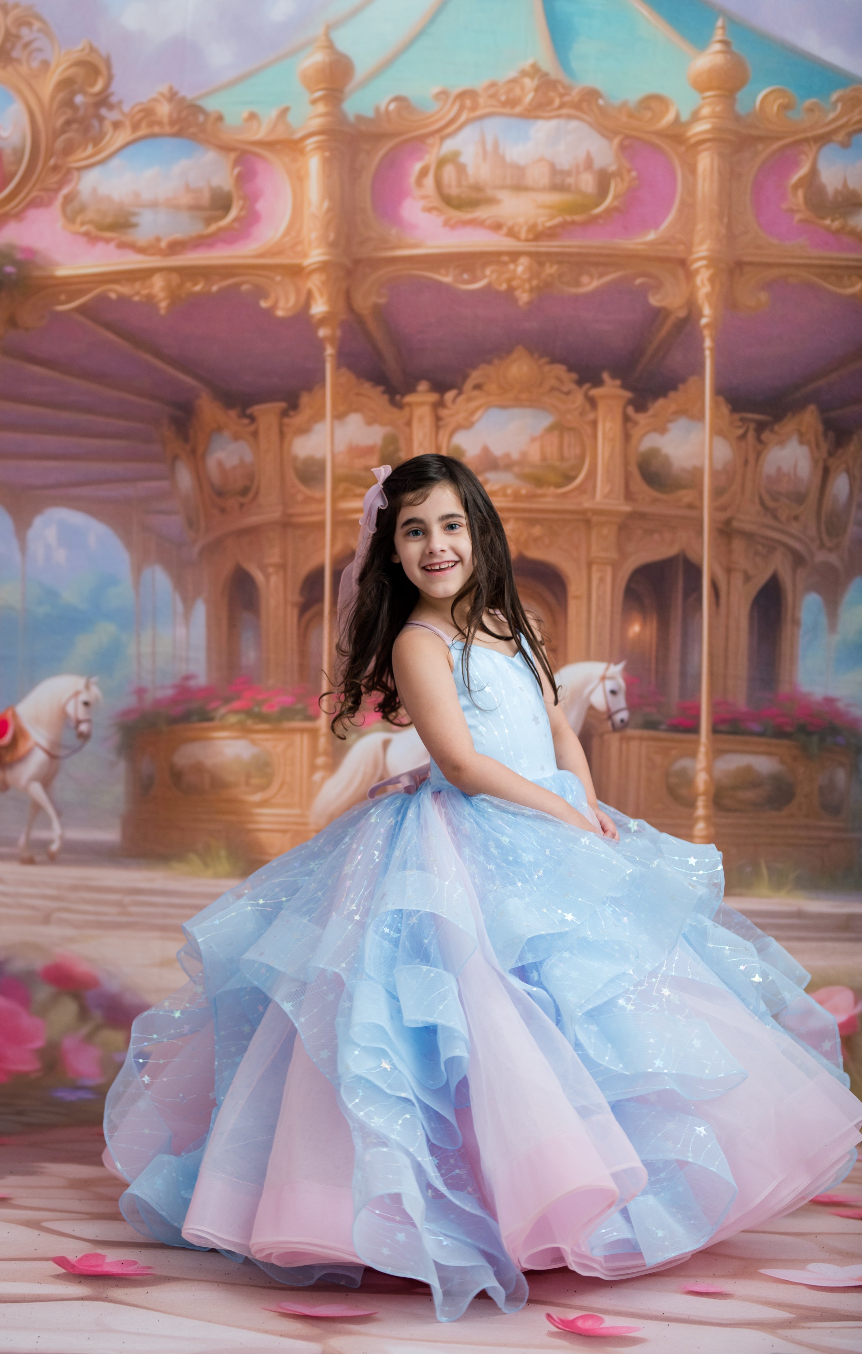 A young girl with long dark hair sits on a carousel, wearing a voluminous light blue and pink ball gown with sparkly details.
