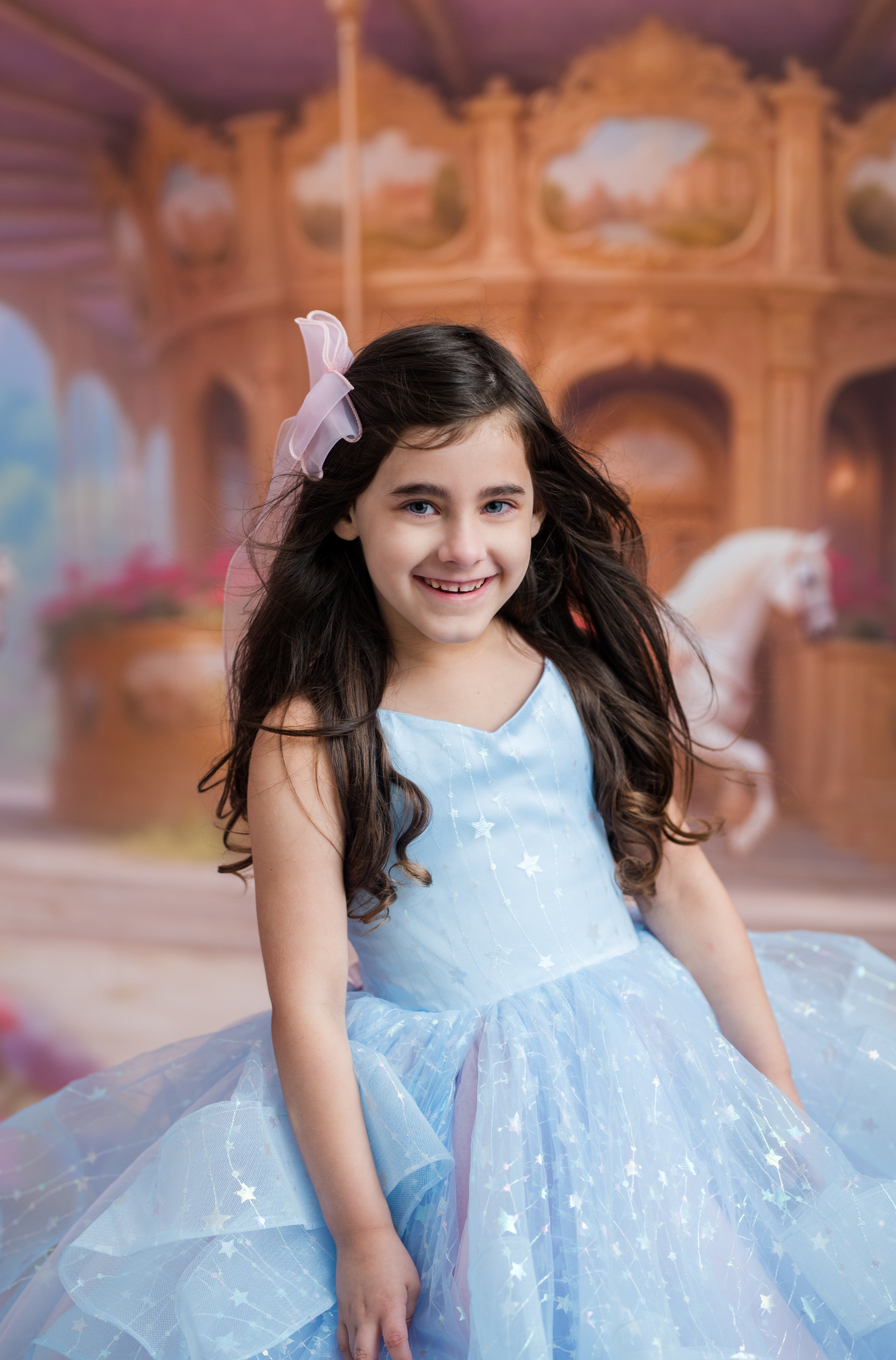 A young girl with long dark hair wearing a light blue sparkly tulle dress and a pink bow, posing in front of a blurred carousel backdrop.