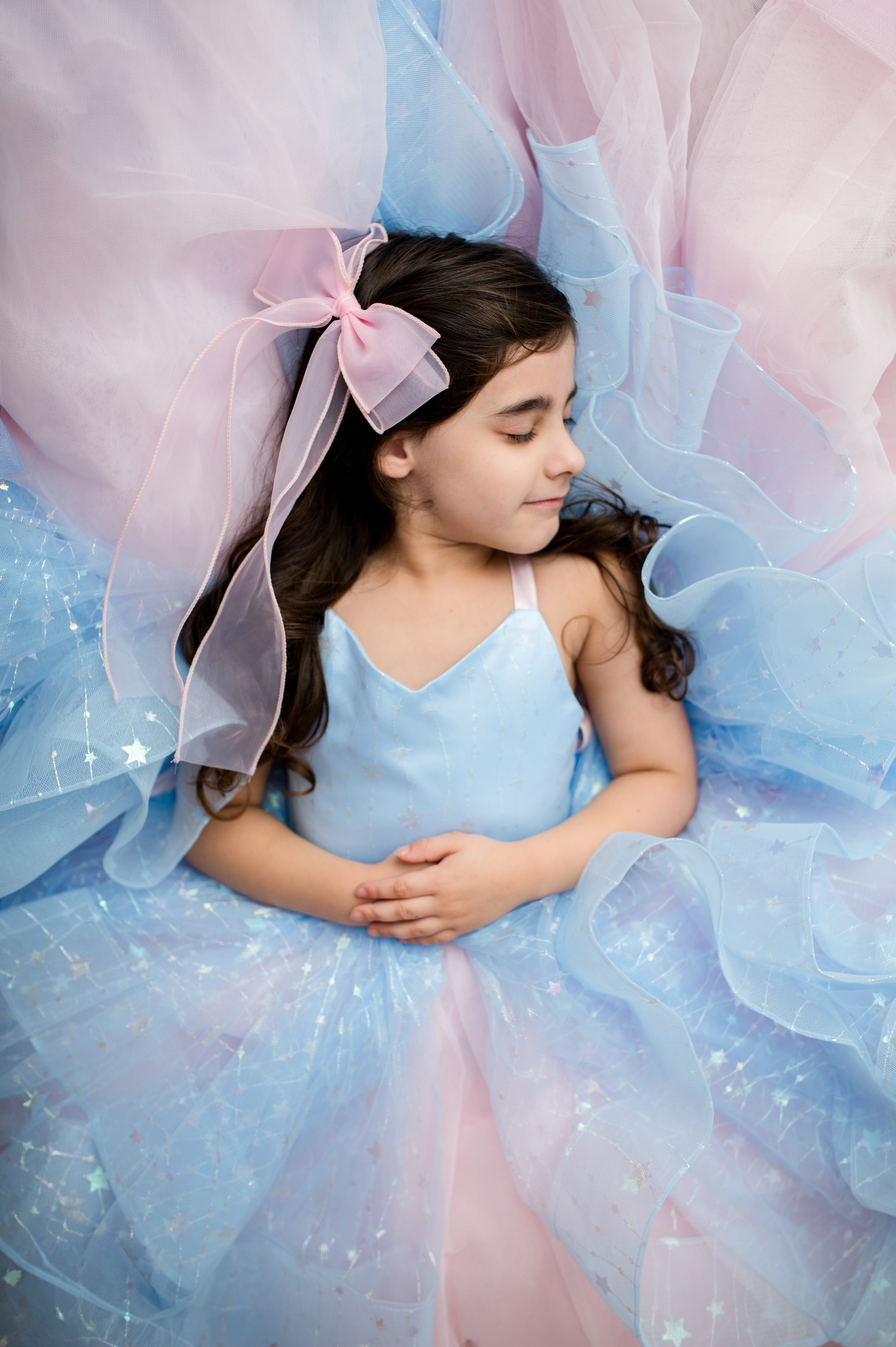 A young girl with long dark hair wearing a light blue spaghetti-strap top and a sparkly pastel blue tulle tutu dress with a pink bow in her hair, posed on a bed of matching pink and blue tulle fabric.