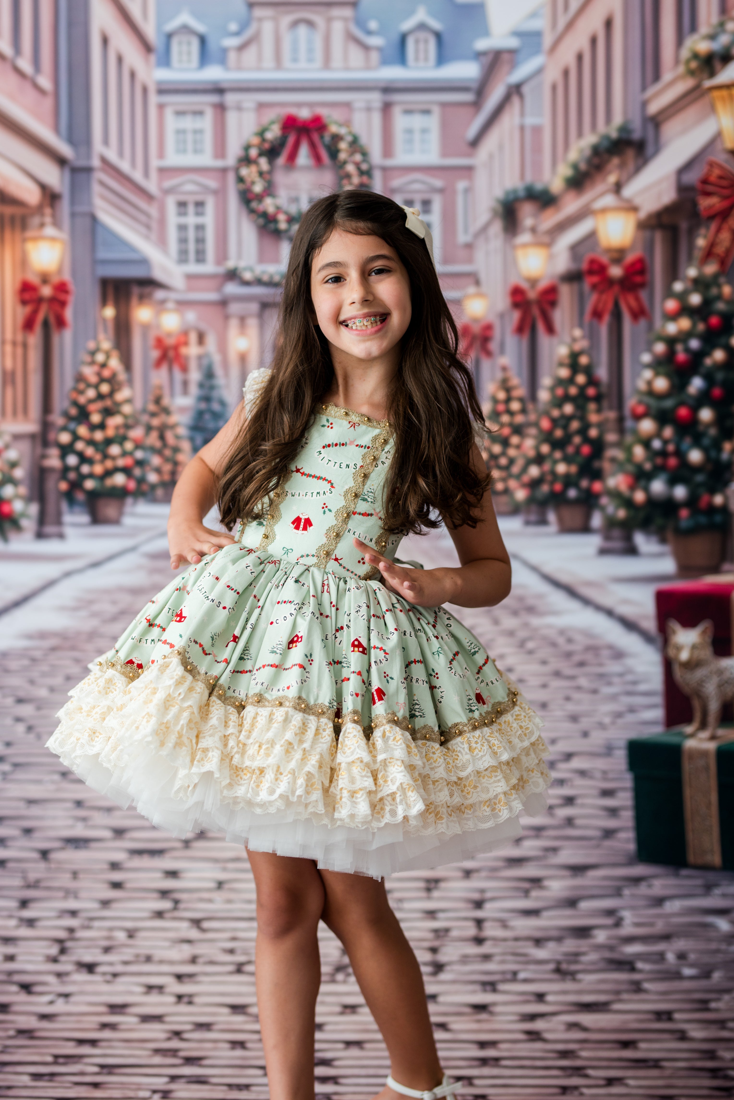 A young girl with long brown hair stands in a festive Christmas-themed street scene wearing a light sage green dress with red and gold holiday motifs and a layered cream ruffle skirt.