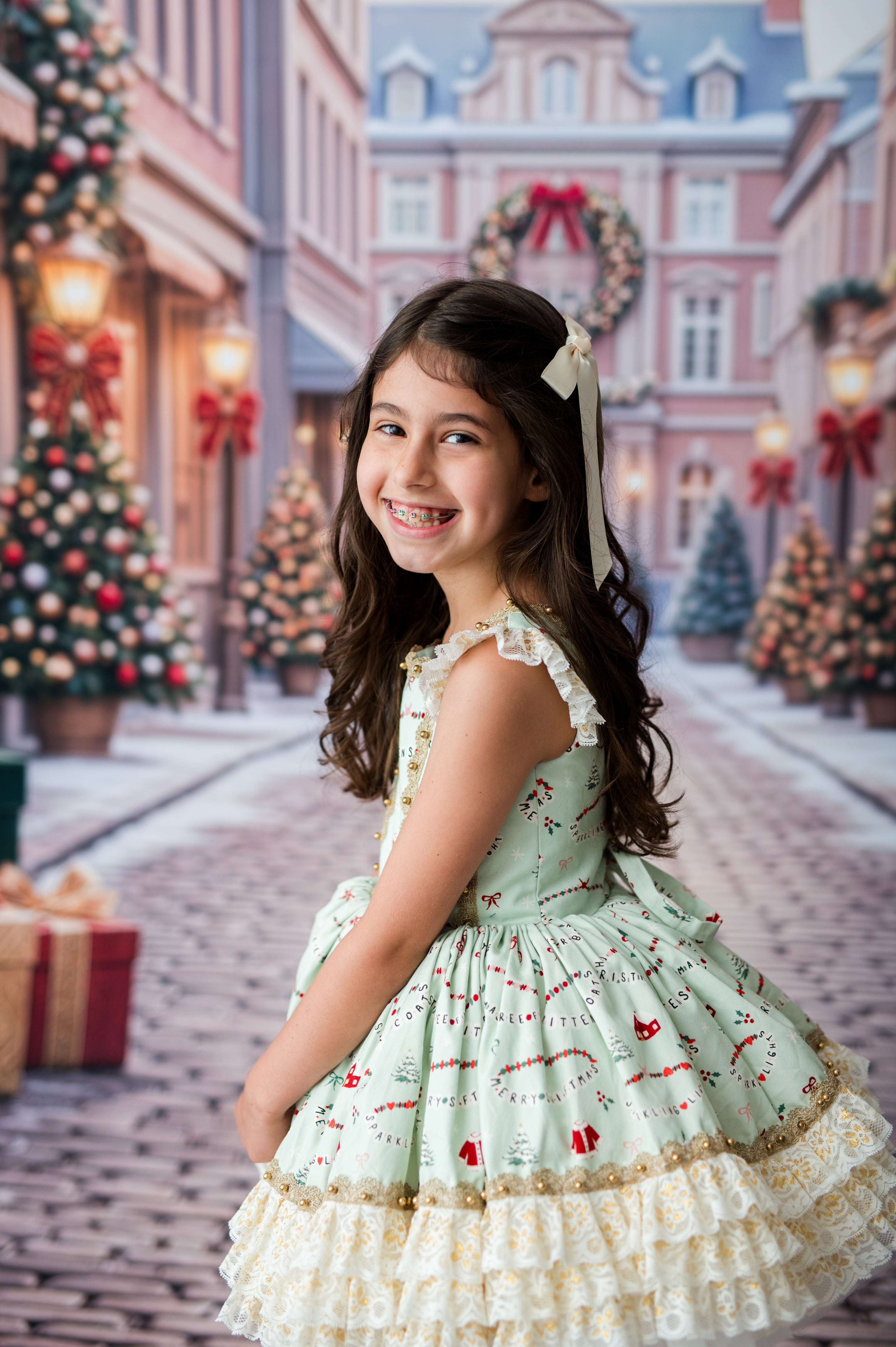 A young girl with long brown hair wearing a light green and cream ruffled Christmas dress, smiling while posing in a festive, snow-covered town square with decorated Christmas trees and buildings.