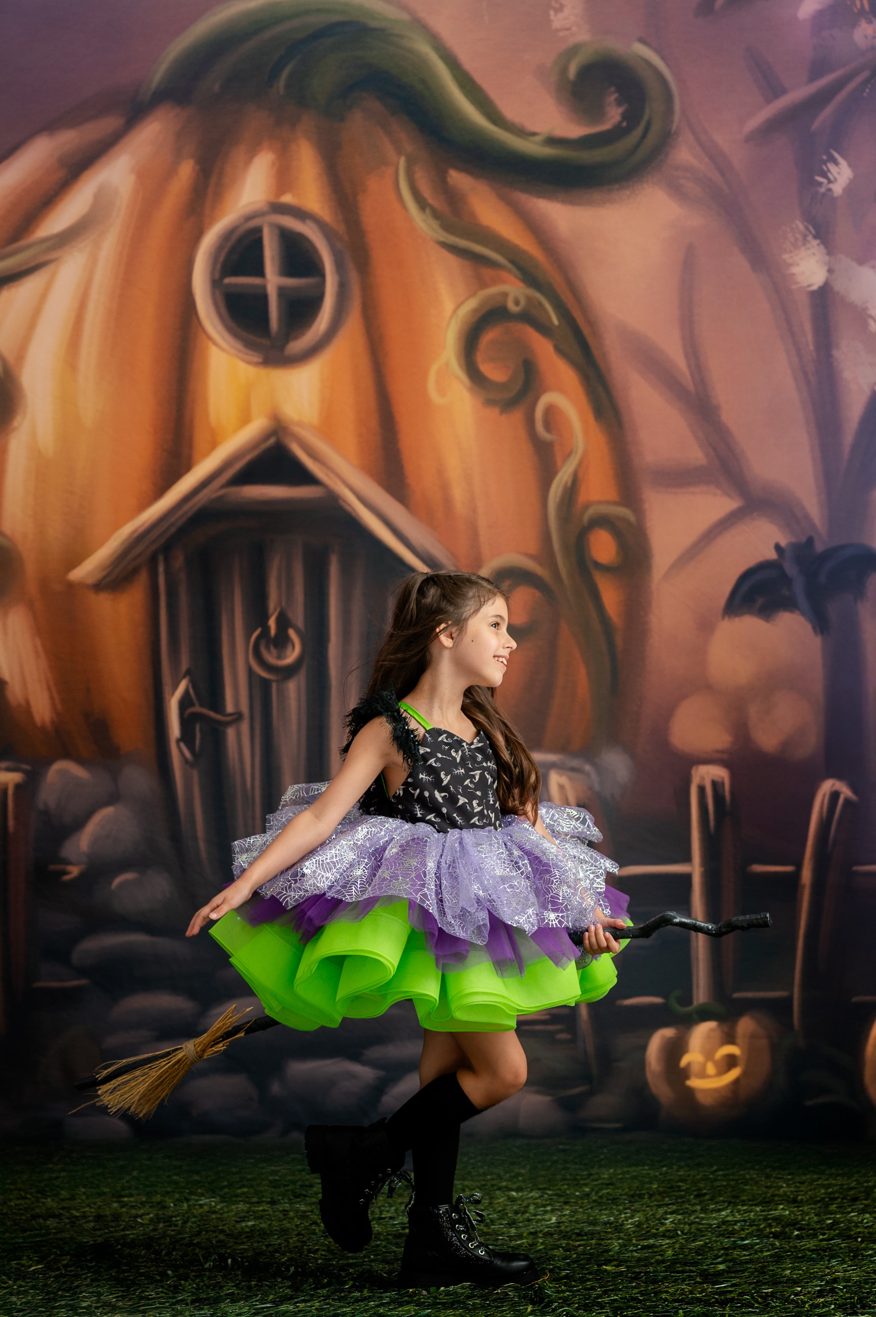 Young girl in a Halloween costume standing in front of a pumpkin house backdrop.