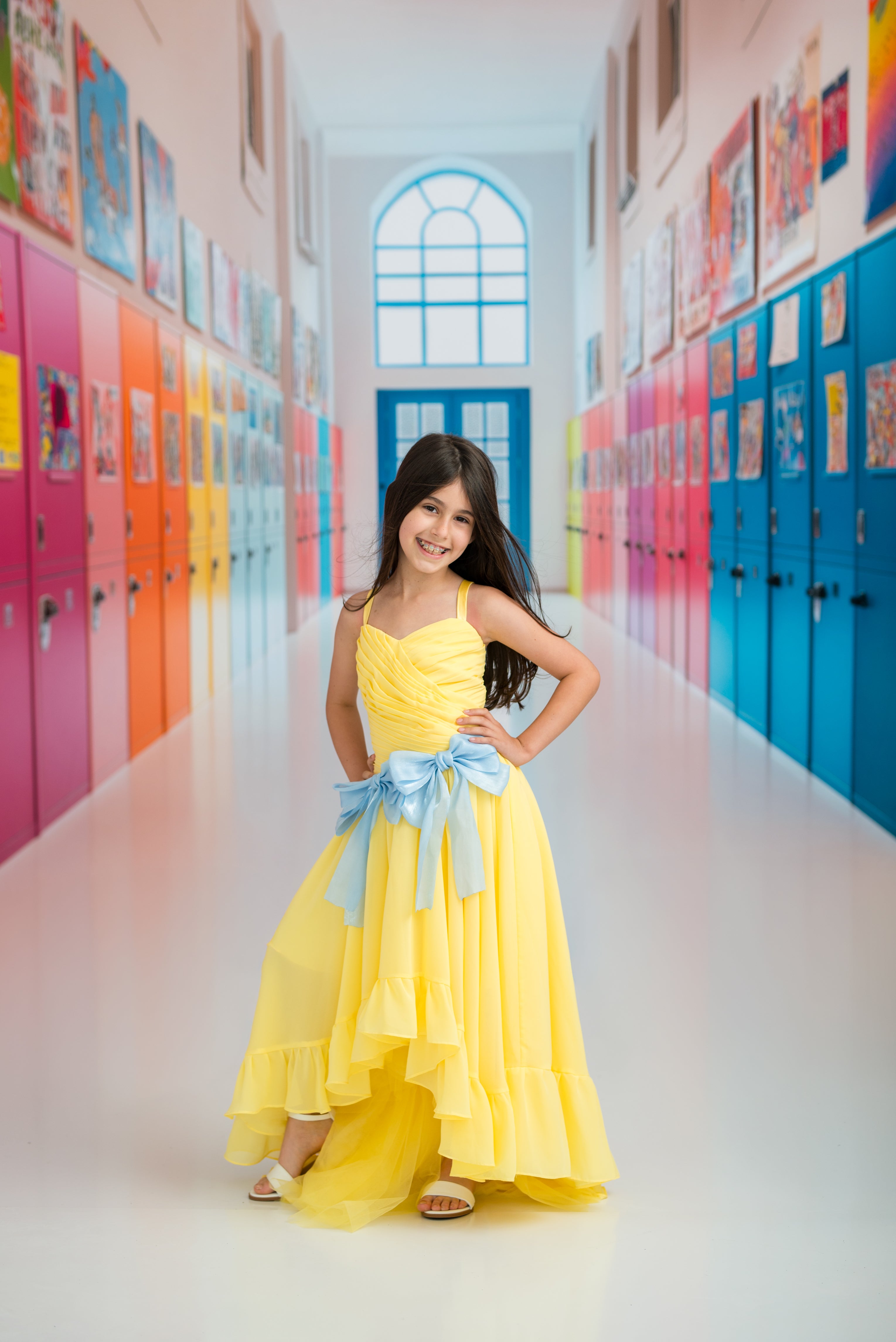 Young girl in a yellow dress with a blue bow standing in a colorful hallway.