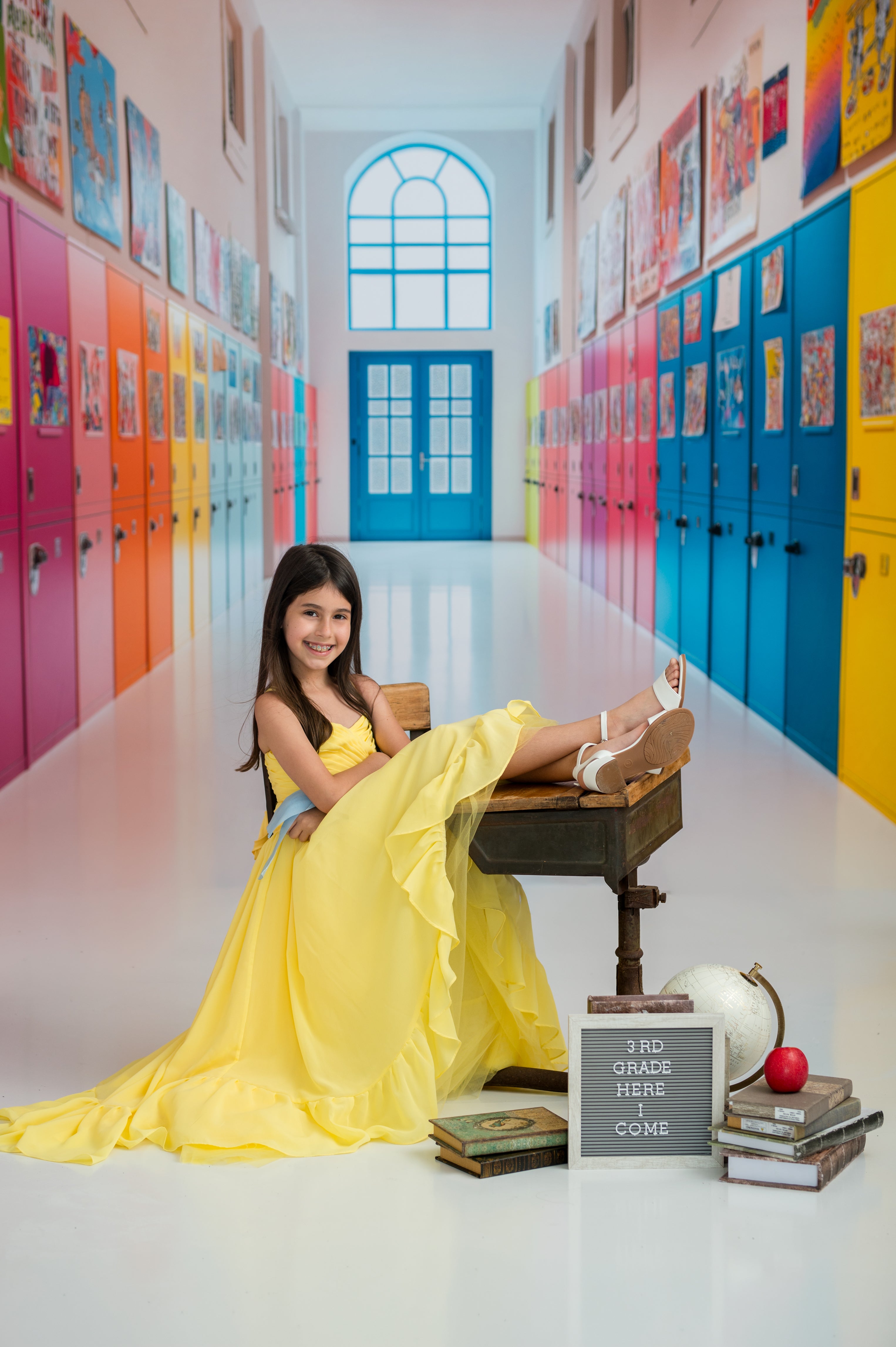 Young girl in a yellow dress sitting on a desk in a colorful school hallway.