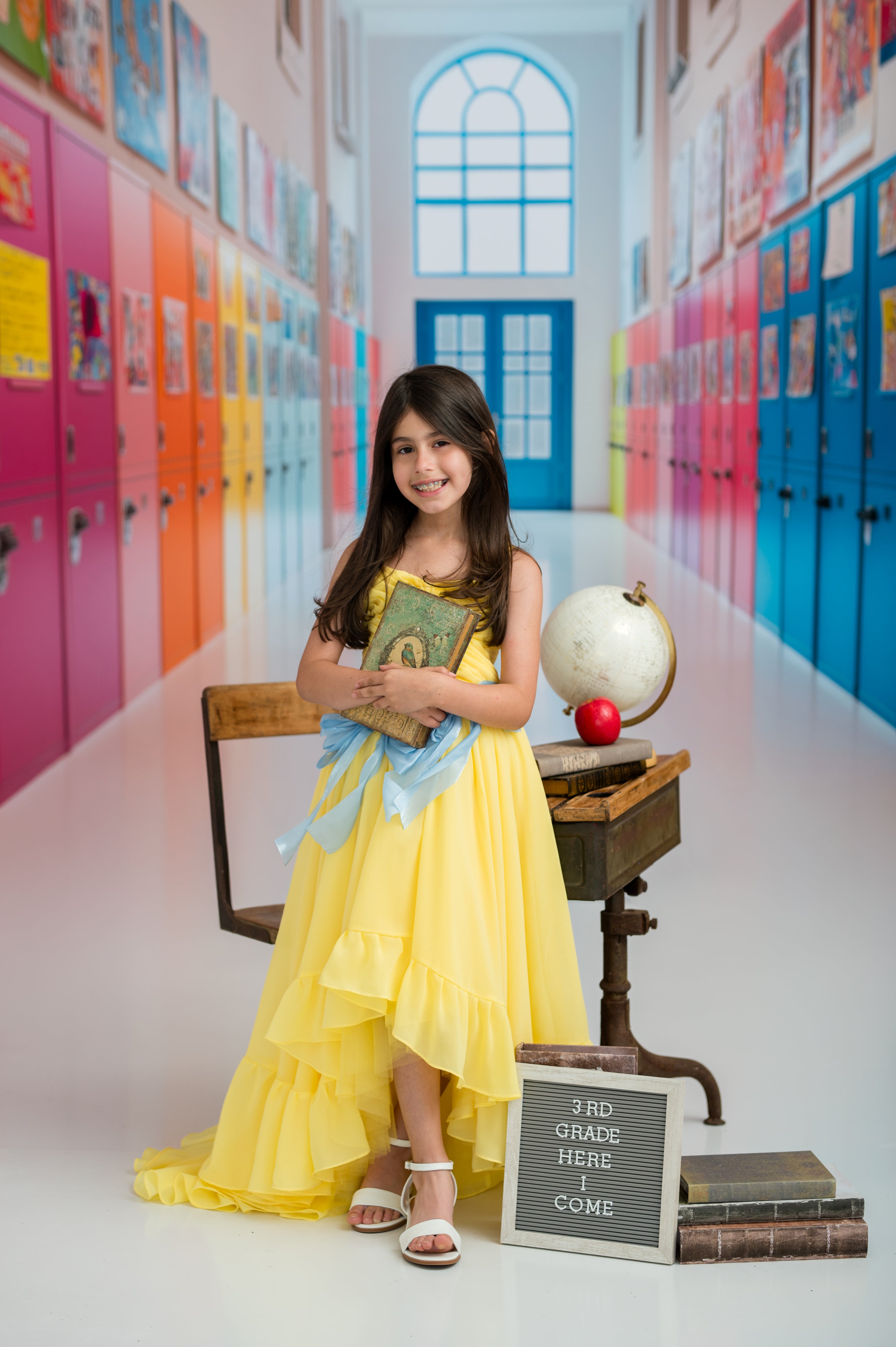 Young girl in a yellow dress sitting in a colorful school hallway with lockers.