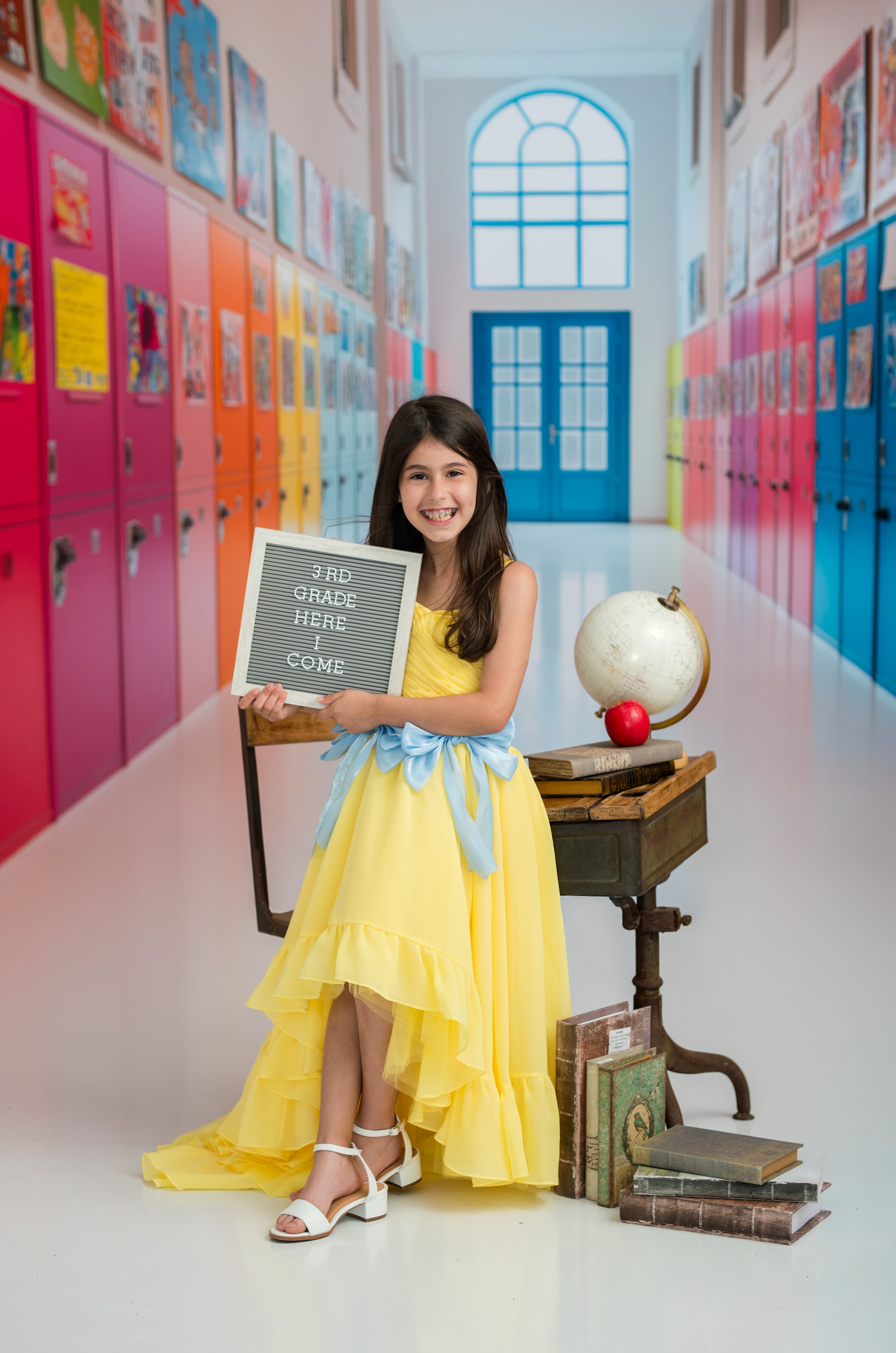 Young girl in a yellow dress holding a sign in a colorful school hallway.