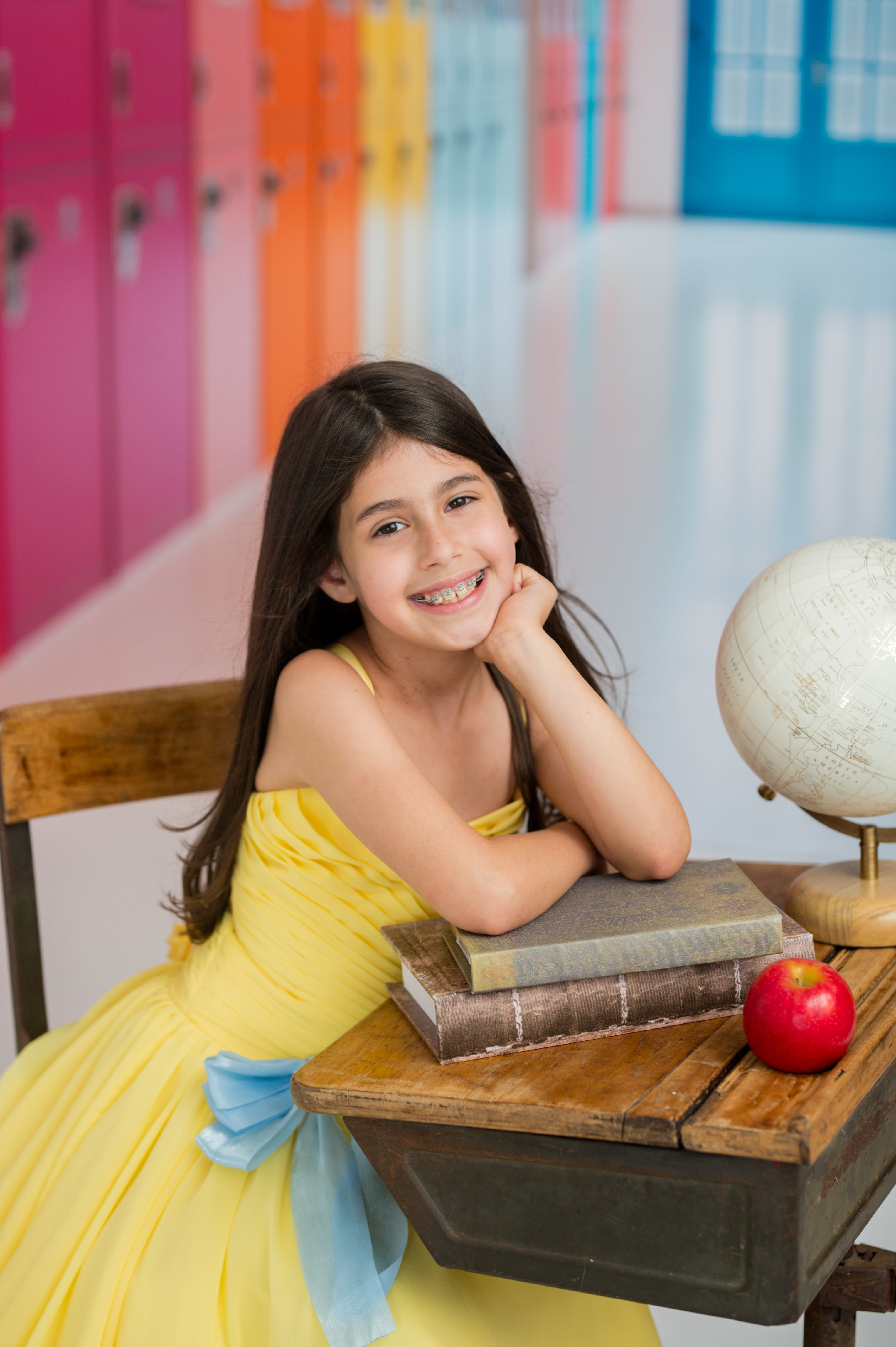 Young girl in a yellow dress sitting at a desk with books and an apple, colorful lockers in the background