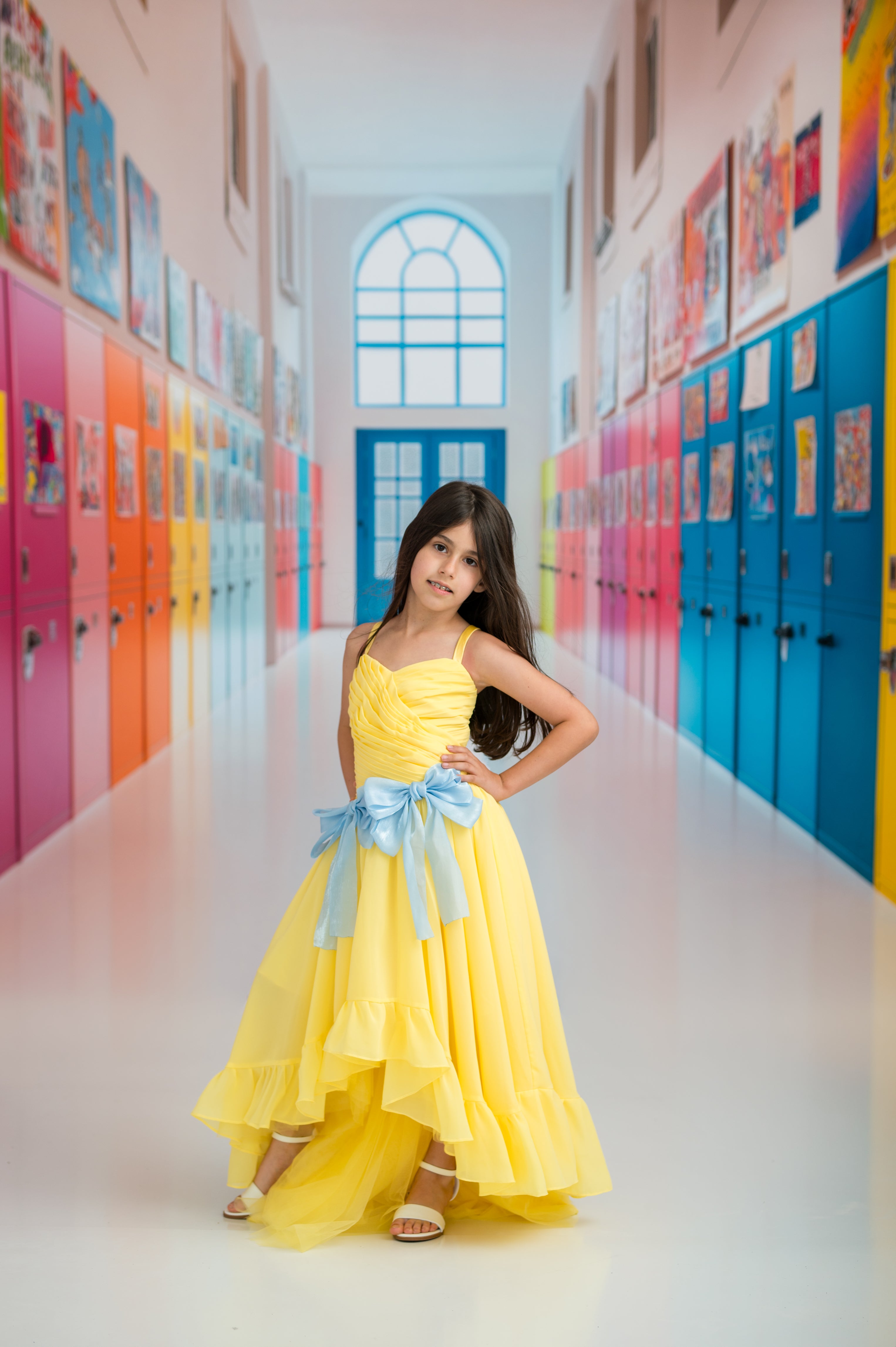 Young girl in a yellow dress with a blue bow standing in a colorful school hallway.