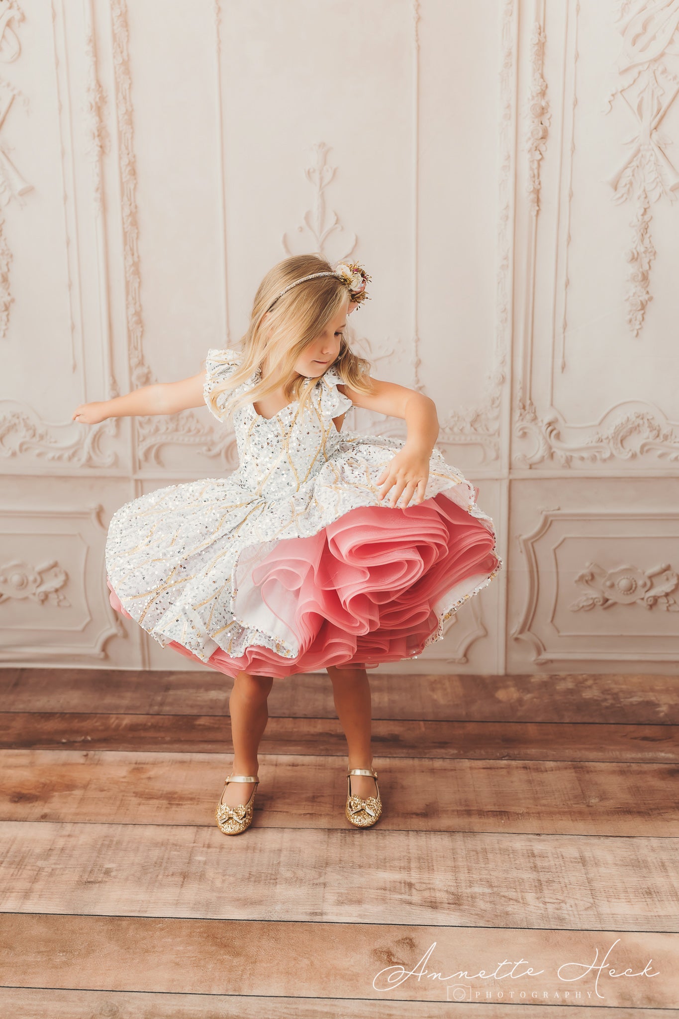 Child wearing a floral headband and a sequined dress with a voluminous pink skirt, gracefully turning on a wooden floor.