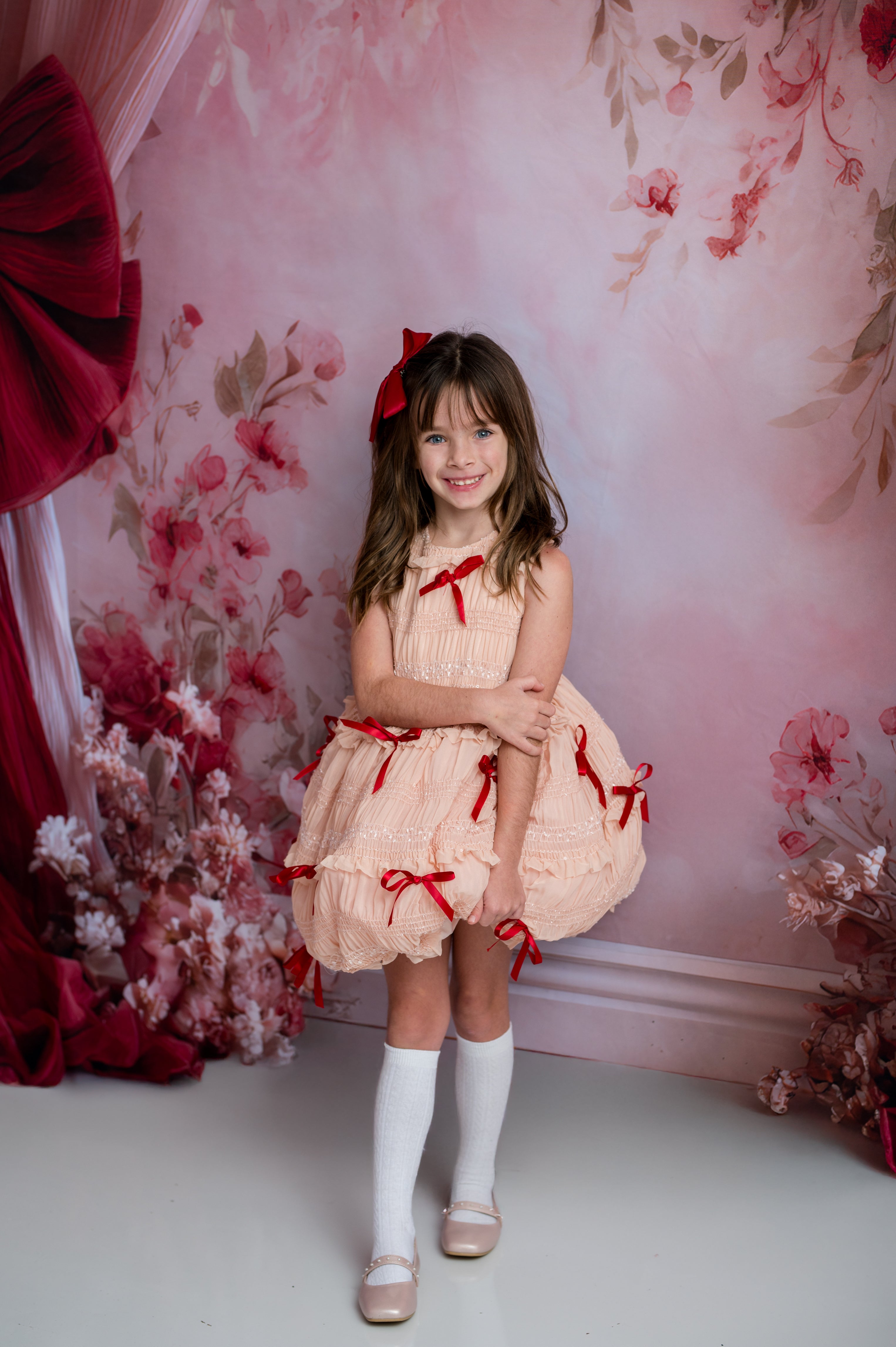 Young girl in a peach dress with red bows standing against a floral backdrop