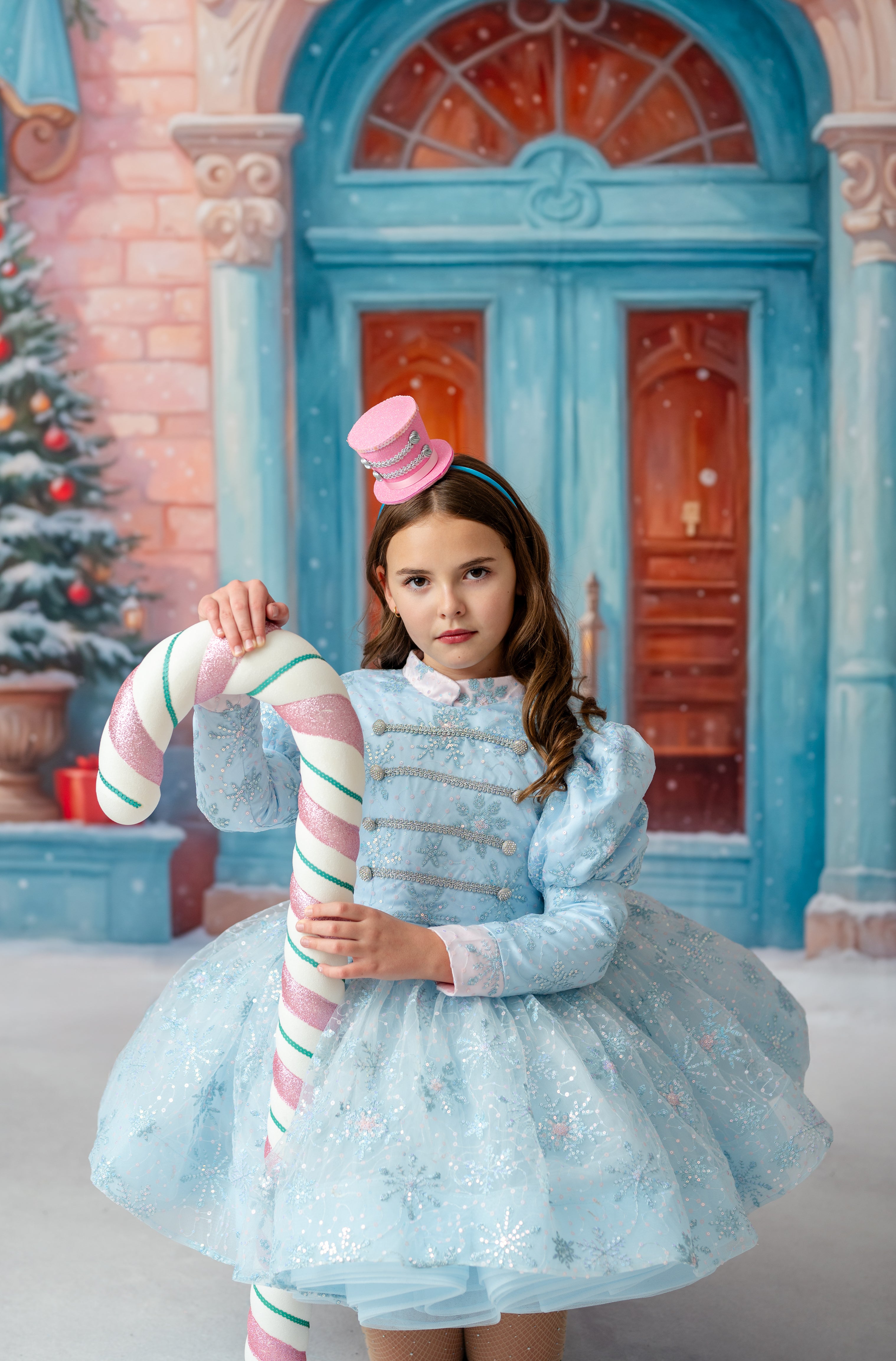 Young girl in a light blue dress holding a candy cane in front of a festive background with a blue door and Christmas tree.