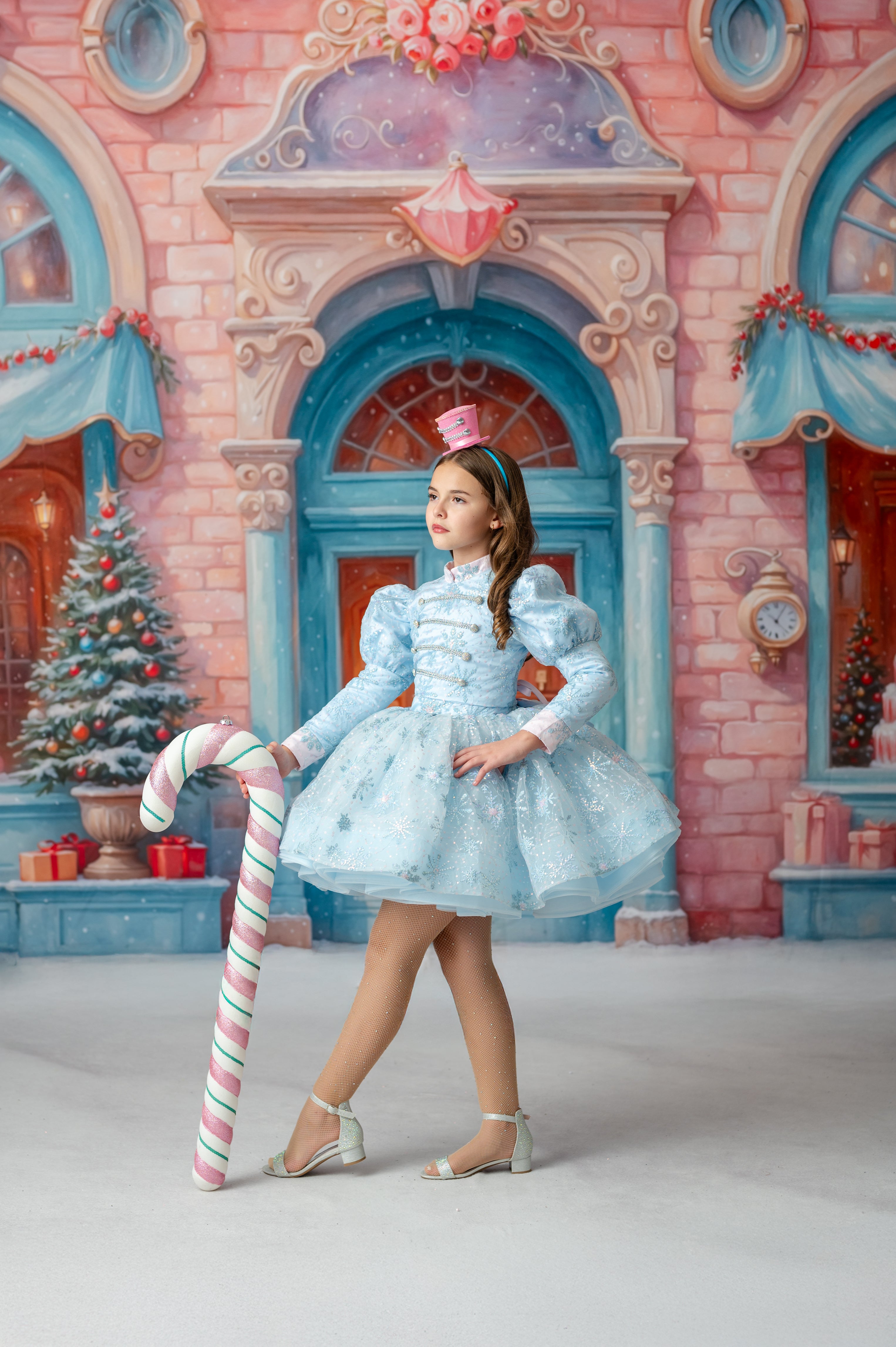 Young girl in a light blue dress with a large candy cane in front of a festive building.