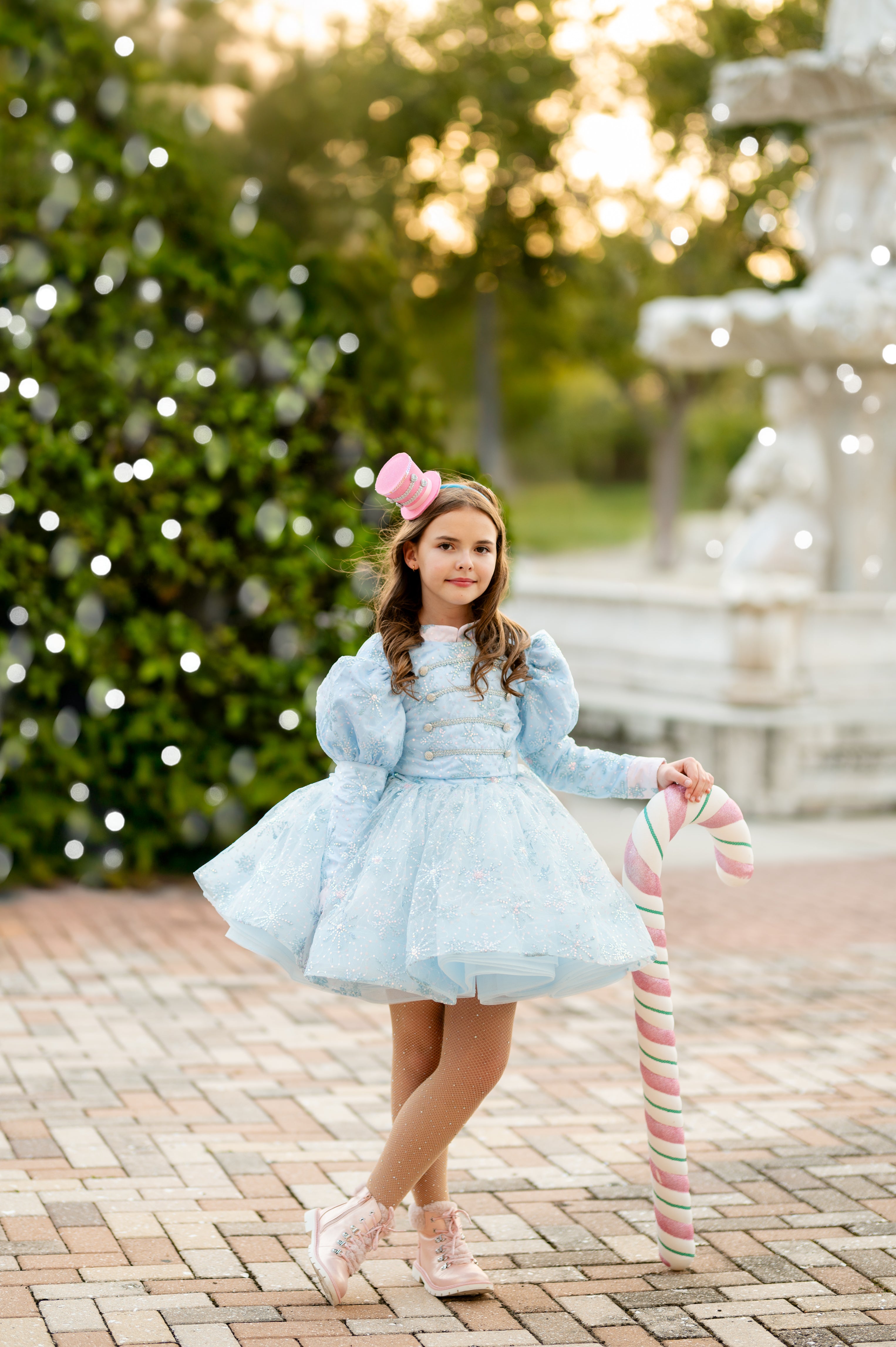 Young girl in a light blue dress with pink bow and candy cane walking outdoors.