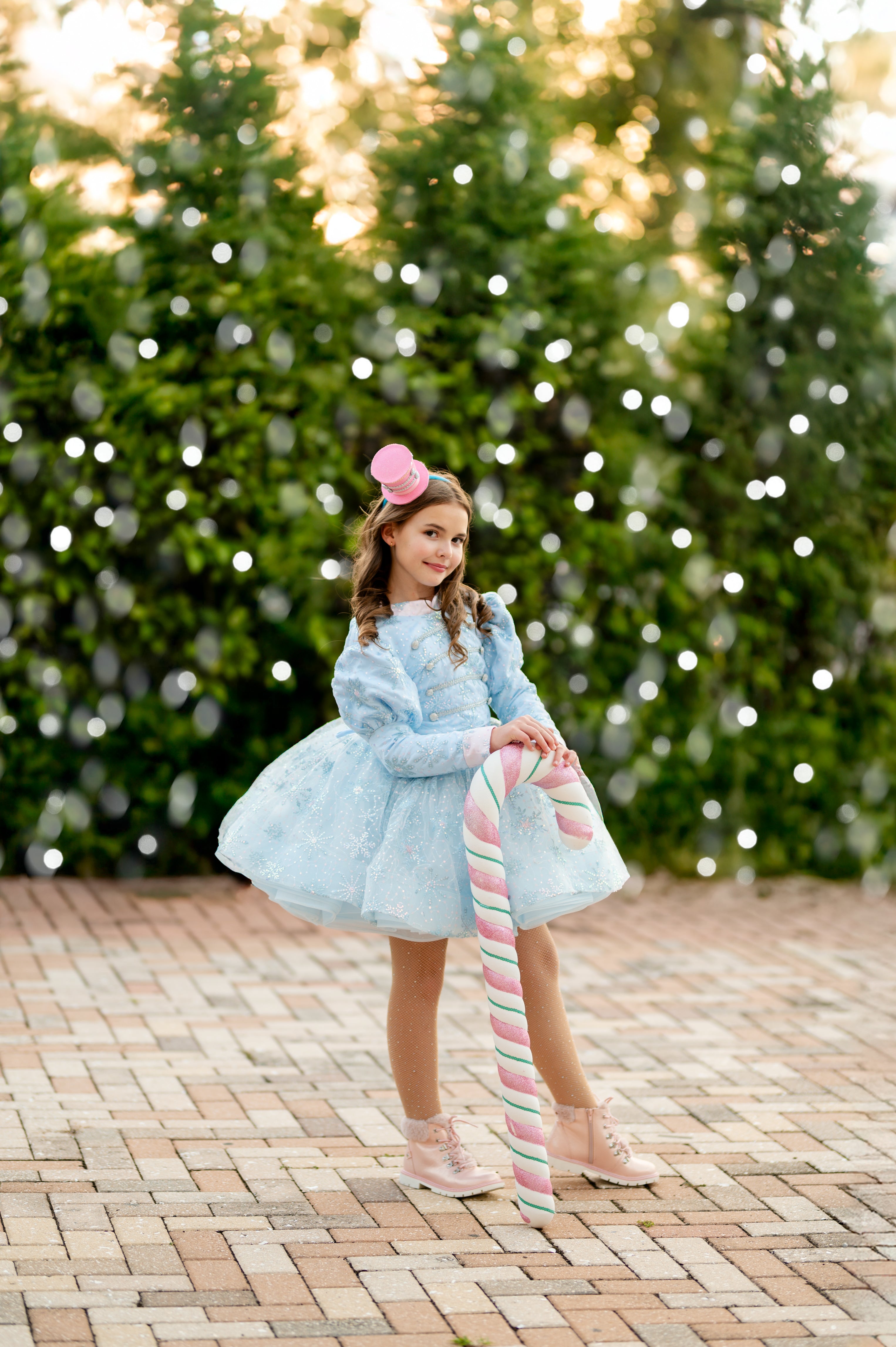 Young girl in a light blue dress holding a striped candy cane against a blurred green and white background.