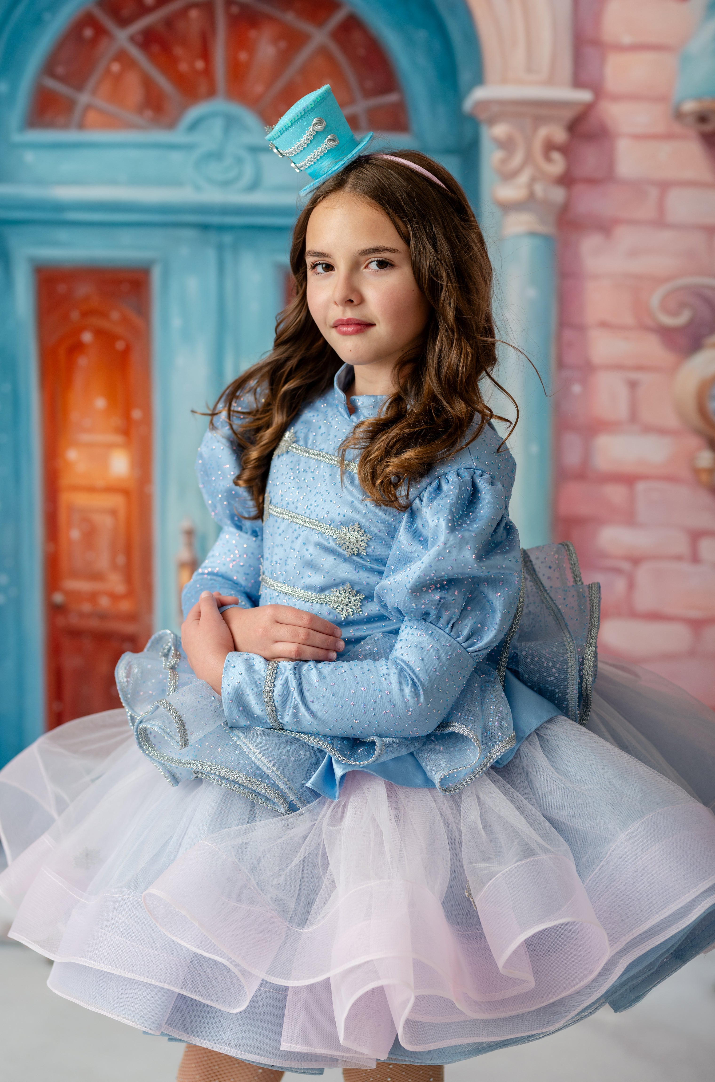 Young girl in a blue and white dress with a hat, standing in front of a colorful castle-like background.