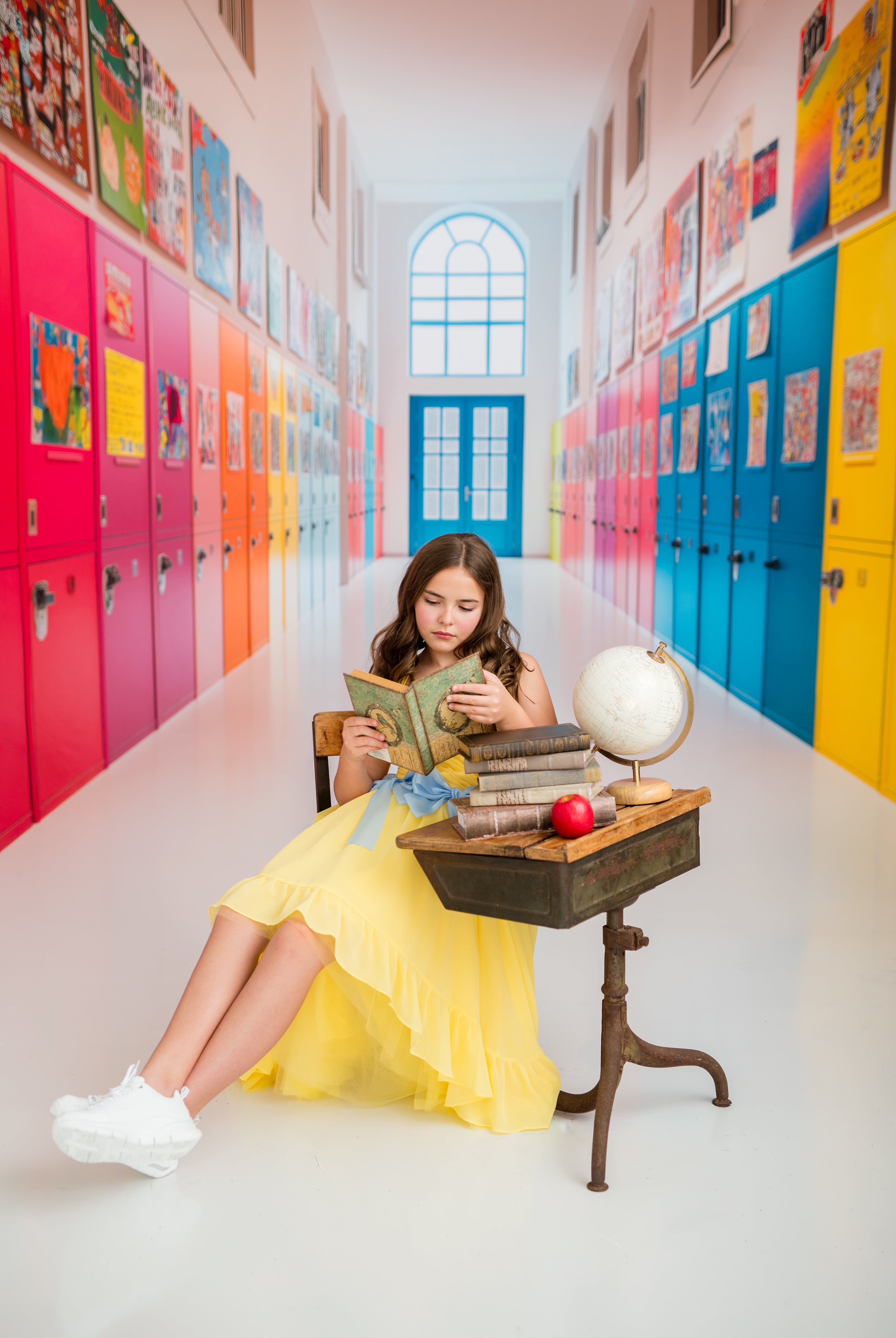 Young girl in a yellow dress sitting on a desk in a colorful hallway with lockers.