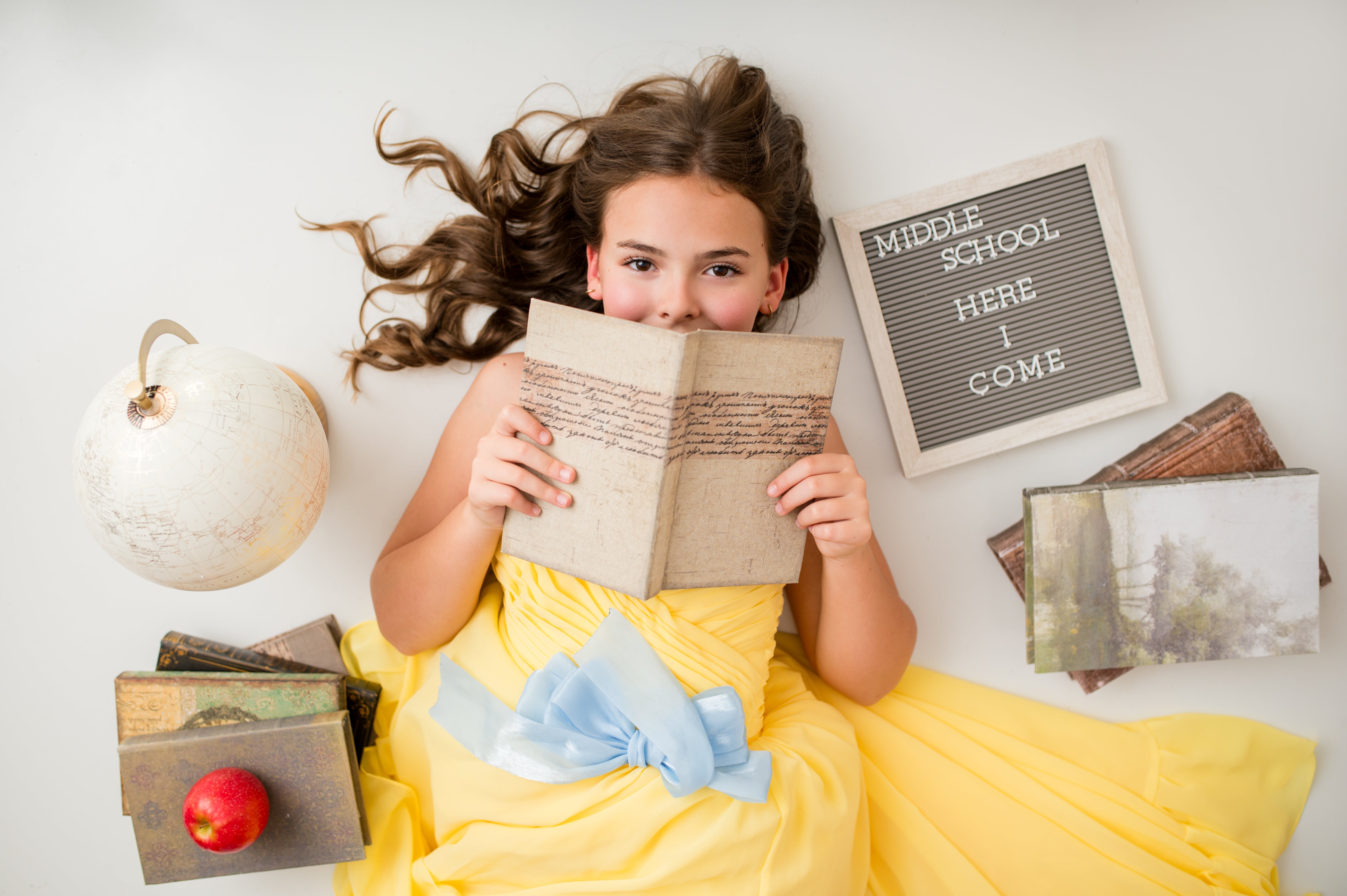 Young girl in a yellow dress holding a book with school-themed decorations around her.
